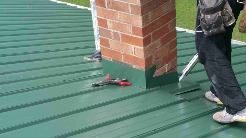 Green metal roof with brick chimney, workers sealing around it with a sealant gun.