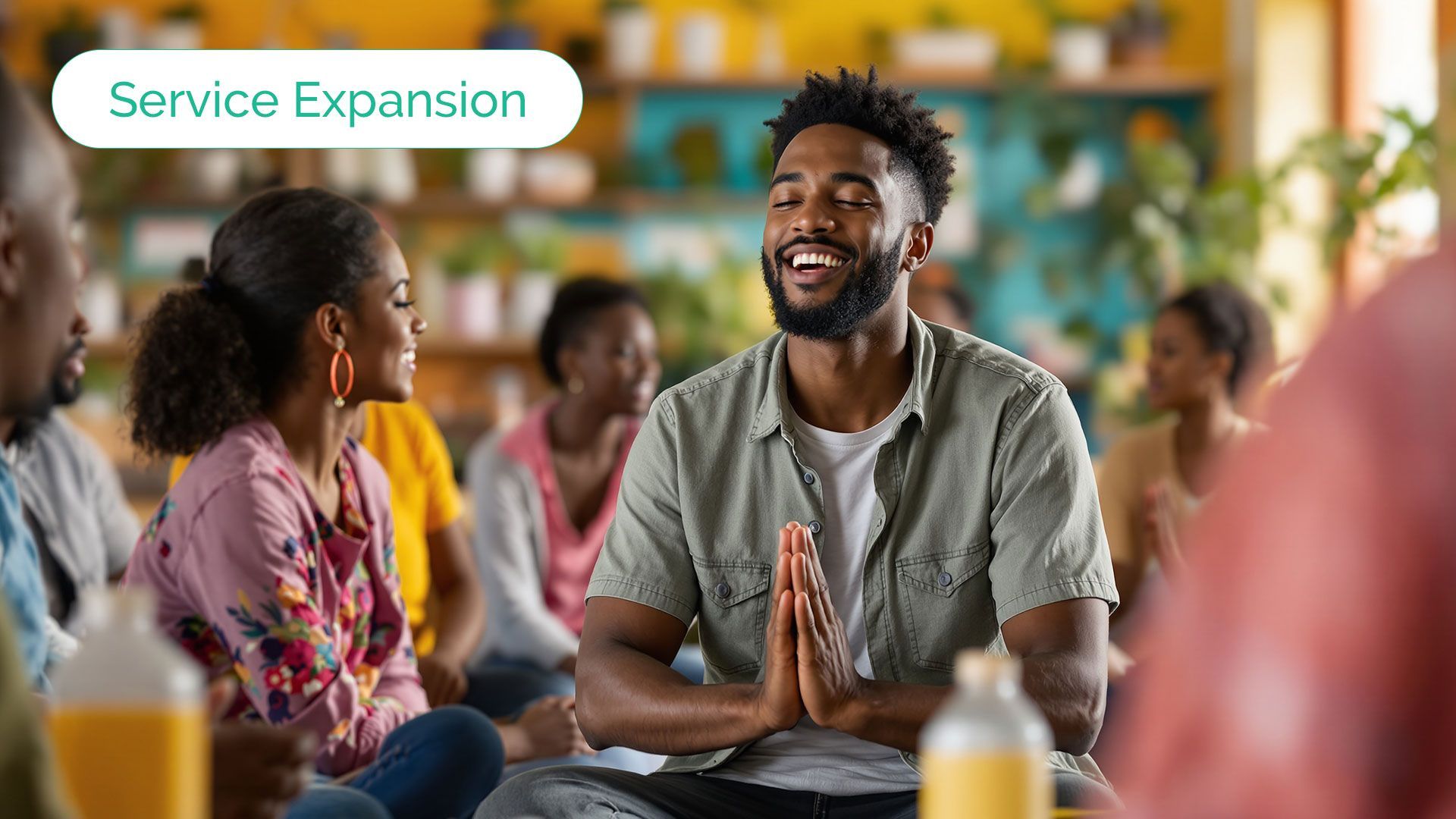 Group of people meditating, smiling, with hands in prayer position. Inside a brightly lit room.