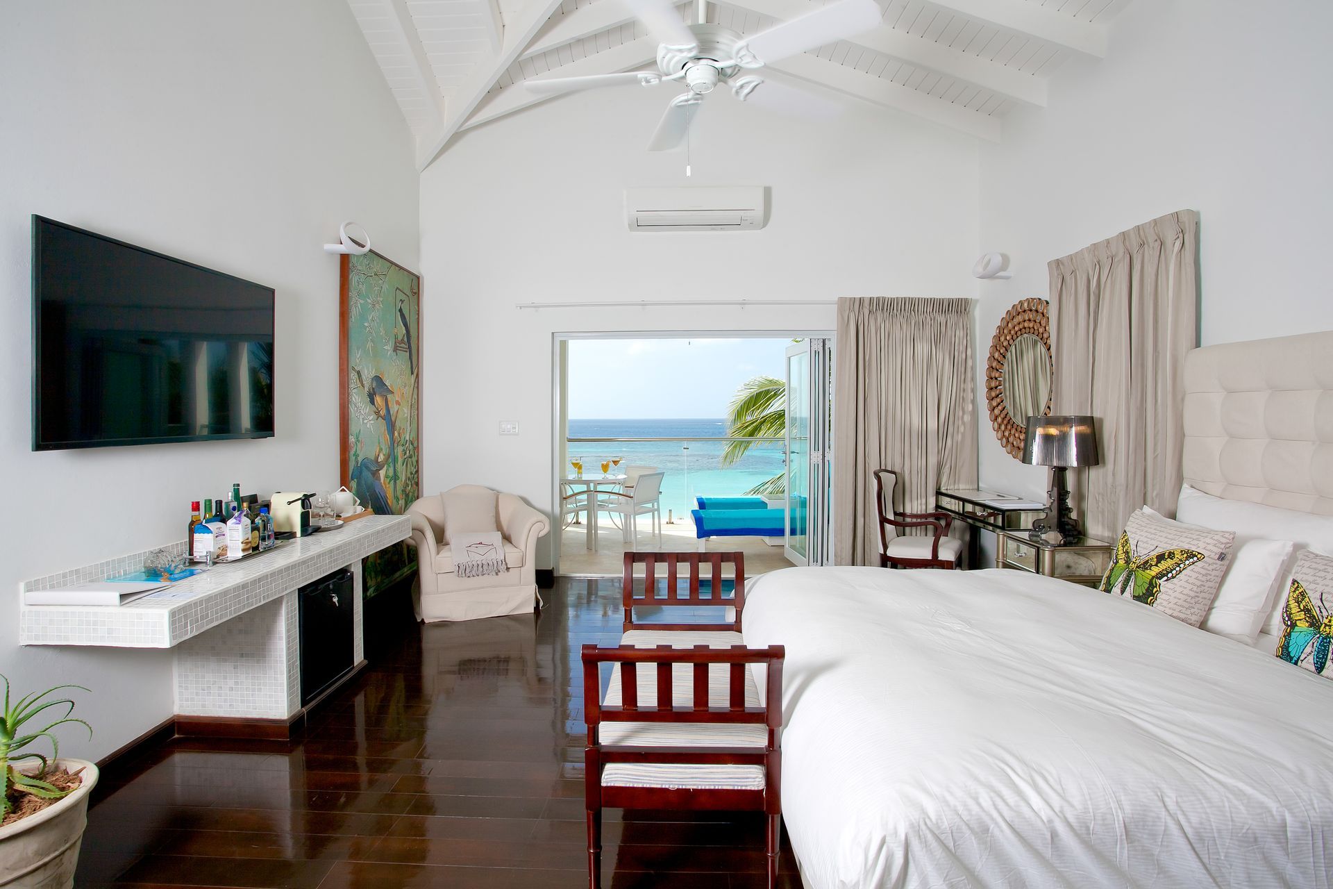 Bright bedroom with ocean view; white walls, dark wood floor, large bed, and open doorway to a patio.