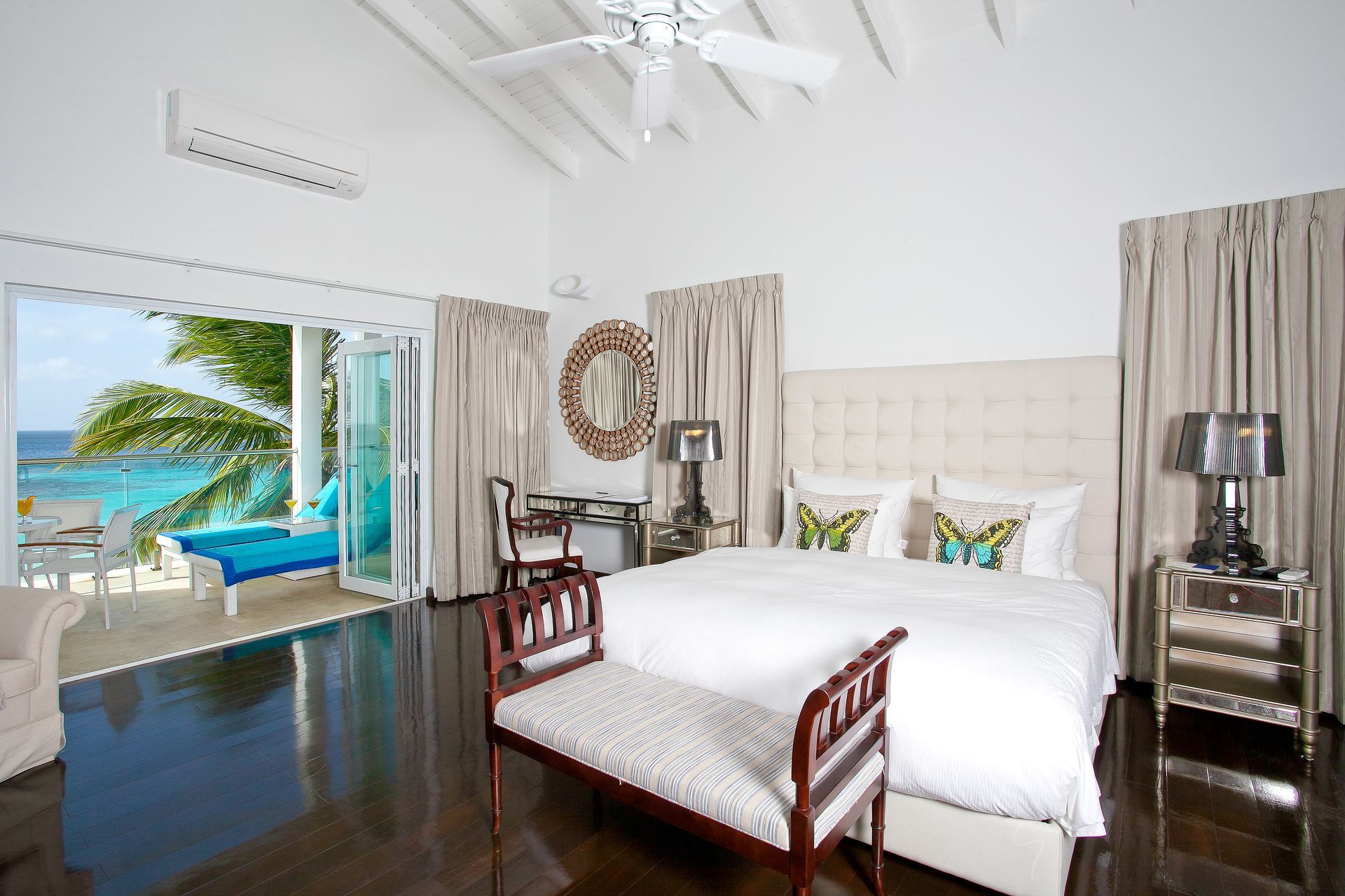 Bedroom with ocean view: white walls, dark wood floor, bed, bench, desk, opening onto a patio.