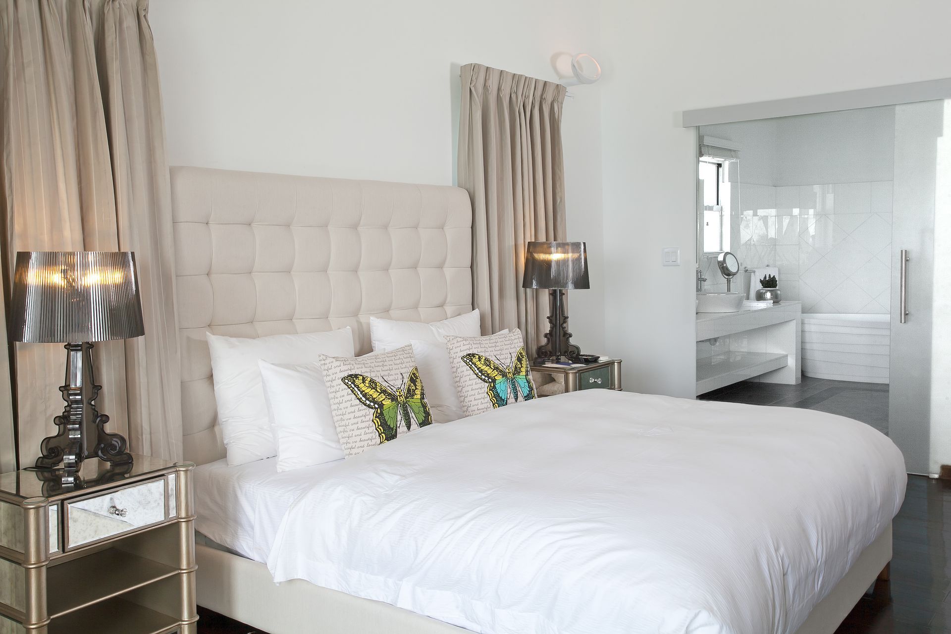 Bedroom with a white bed, mirrored nightstands, neutral curtains, and a sliding glass door to a bathroom.
