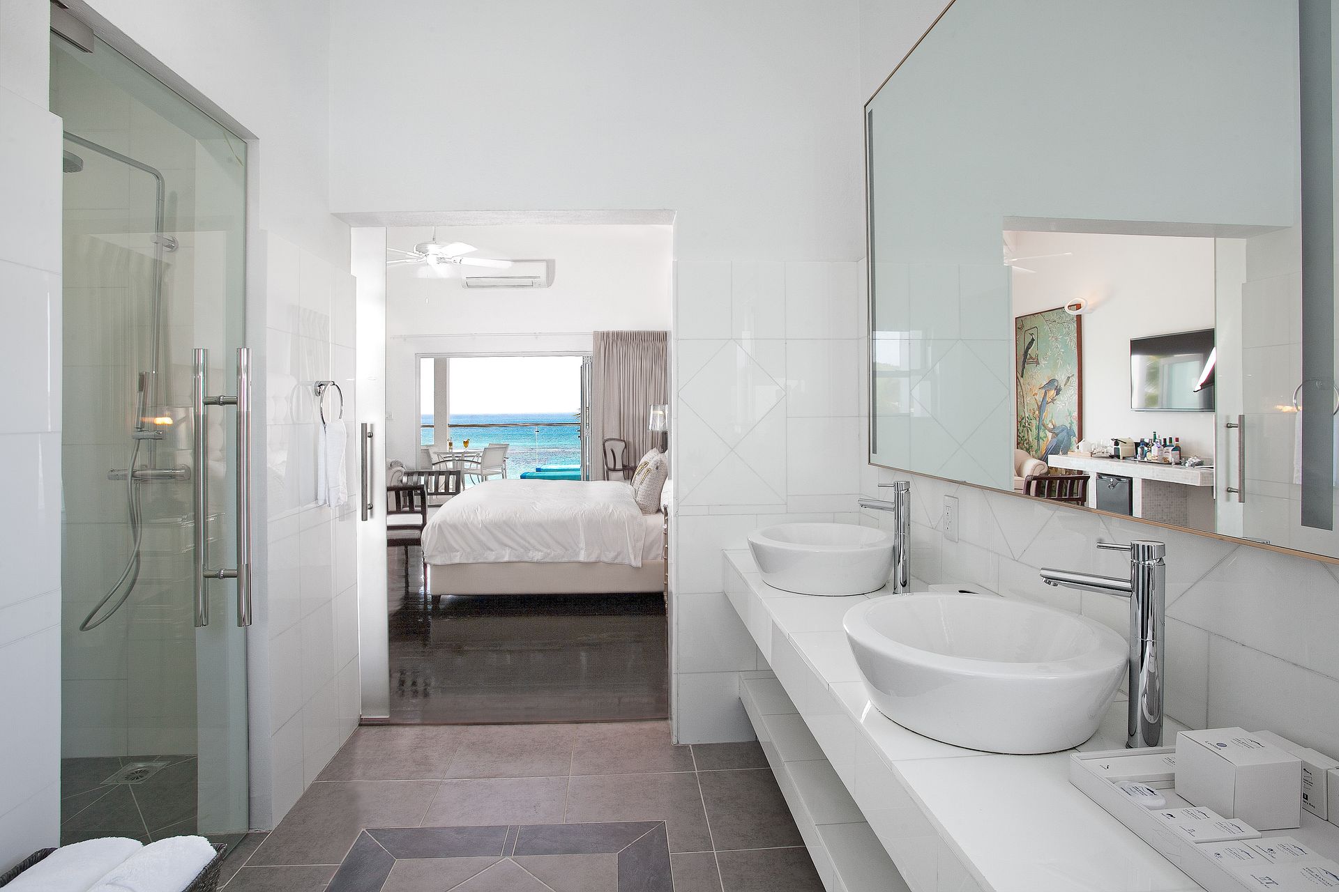 Bright white bathroom with a view into a bedroom with ocean view. Includes a shower, sinks, and a mirror.