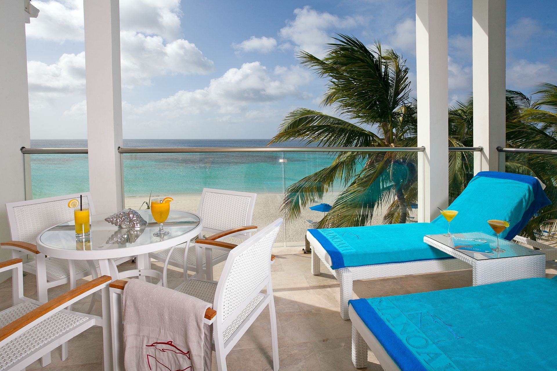 Balcony overlooking a turquoise ocean with lounge chairs, table, and drinks; palm tree in background.