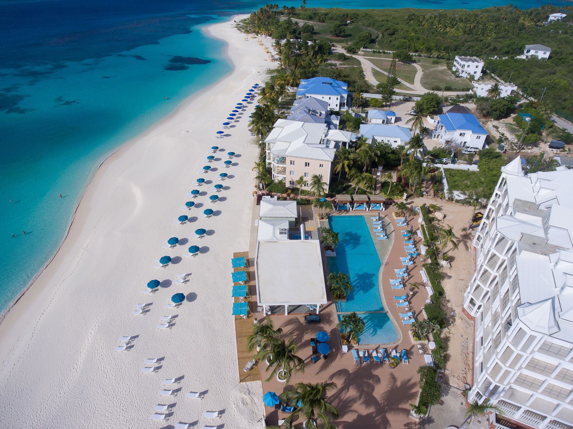 Aerial view of a beach with white sand, turquoise water, buildings, a pool, and blue umbrellas.