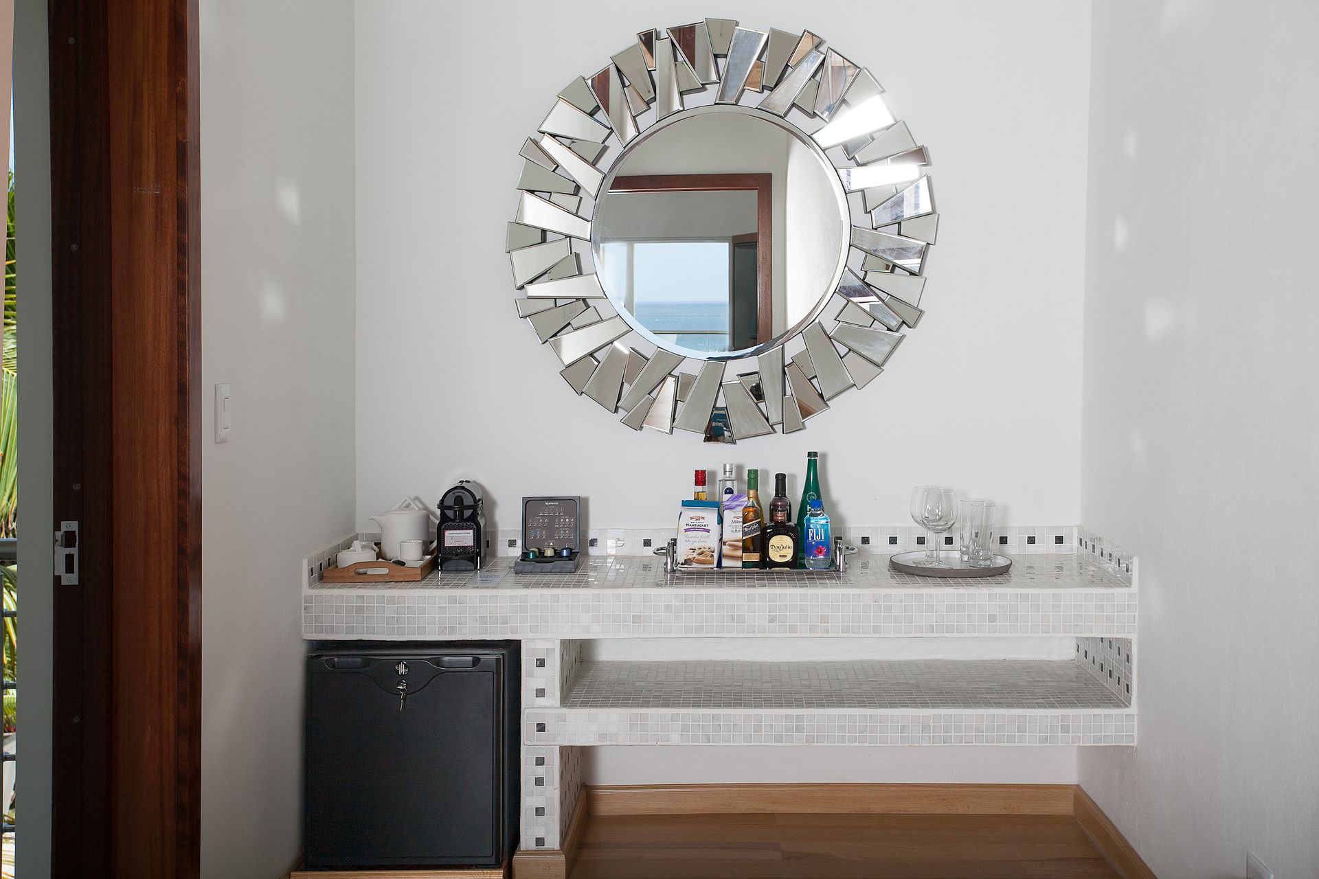 A white wall with a decorative mirror, small bar, and mini-fridge.