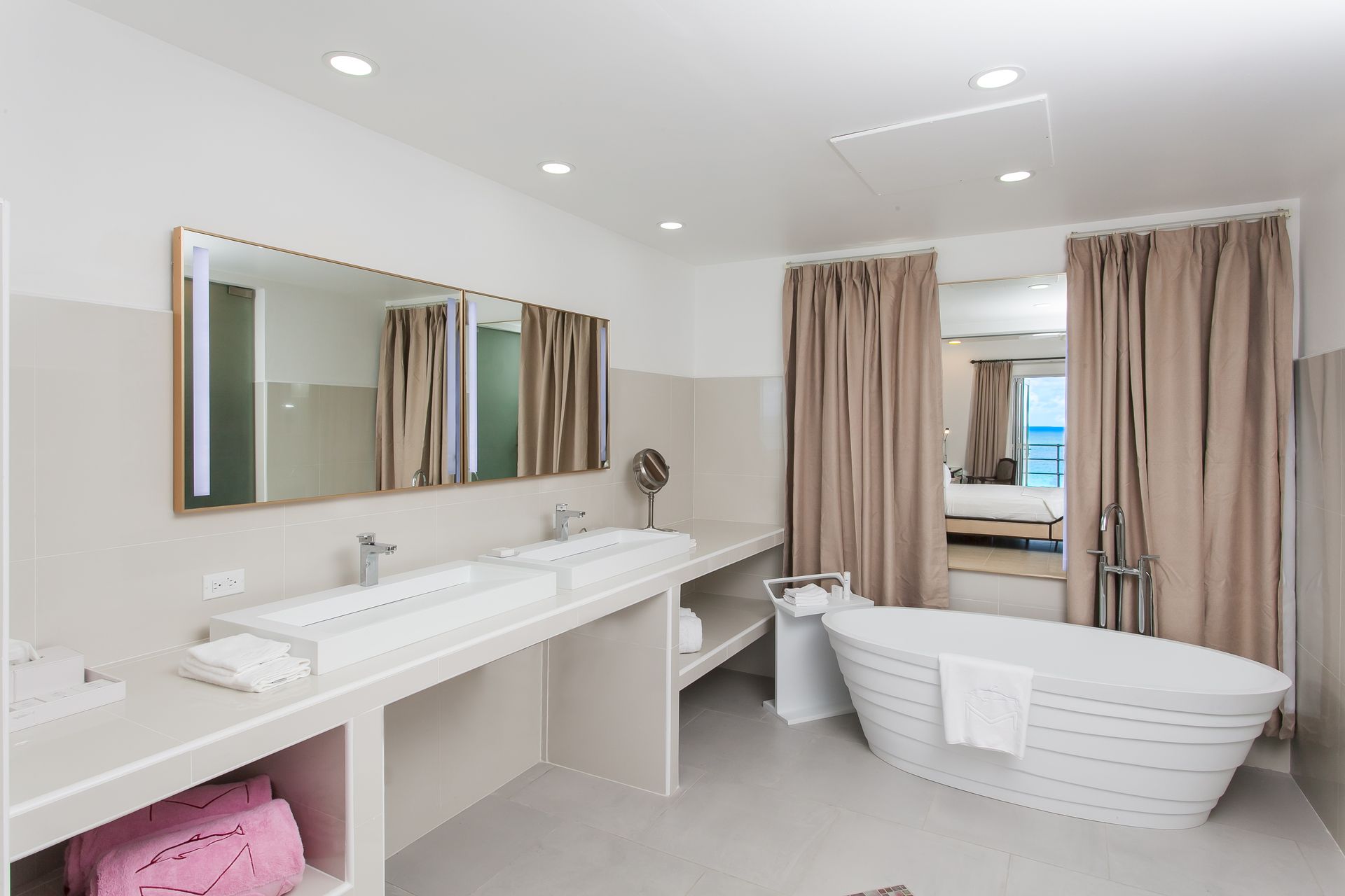 Modern bathroom with a white tub, dual sinks, and large mirrors. Beige curtains frame a view of a bed.