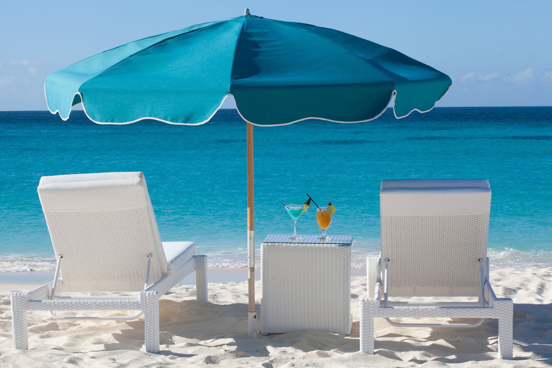 Two white lounge chairs under turquoise umbrella on a sandy beach, cocktails on table, ocean in background.