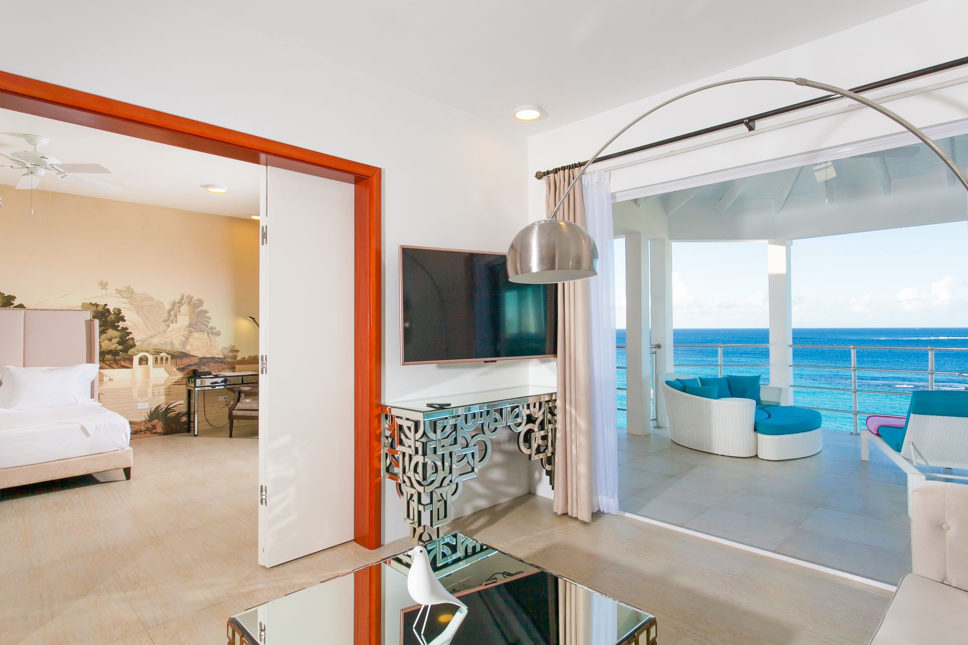 Open-air living room with ocean view, white and teal furniture, bedroom with floral mural visible.