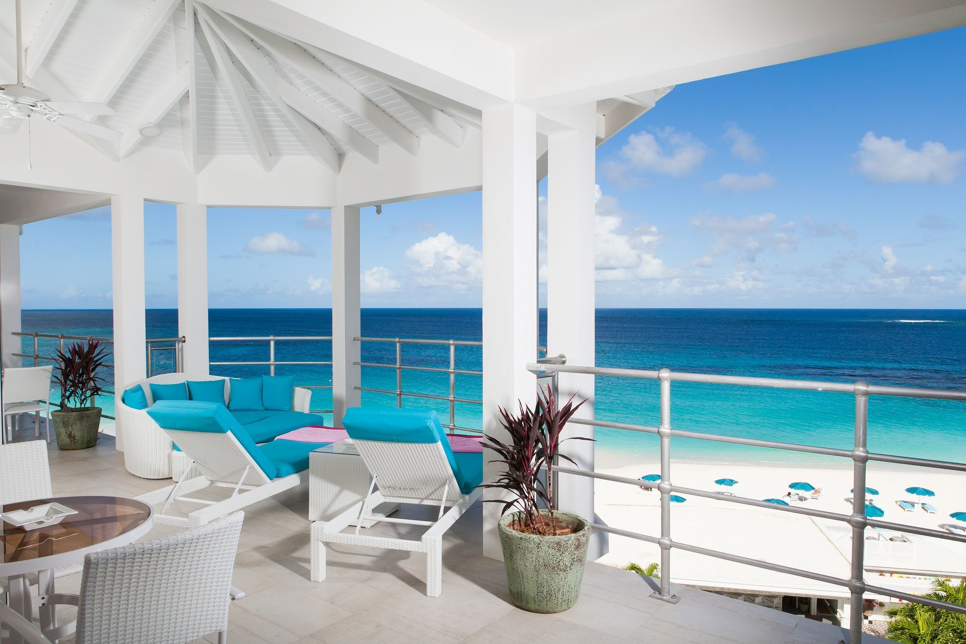 Bright, open-air balcony overlooking a turquoise ocean and white sand beach. Blue seating, white decor.