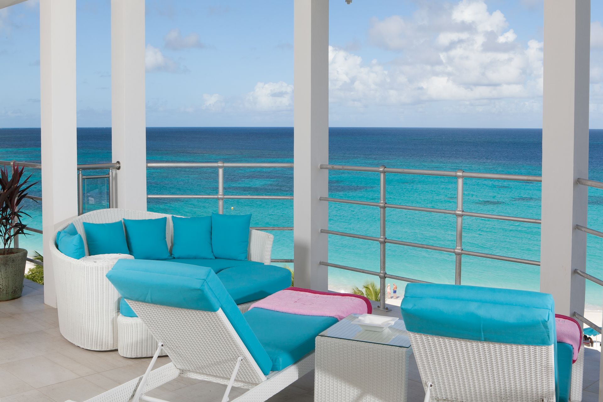 Balcony with white furniture, turquoise cushions, overlooking a blue ocean under a partly cloudy sky.