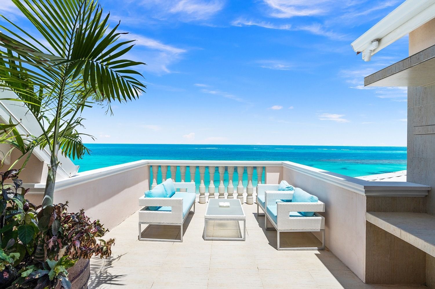 Balcony overlooking turquoise ocean with white furniture and blue cushions. Palm tree on the left. Sunny day.
