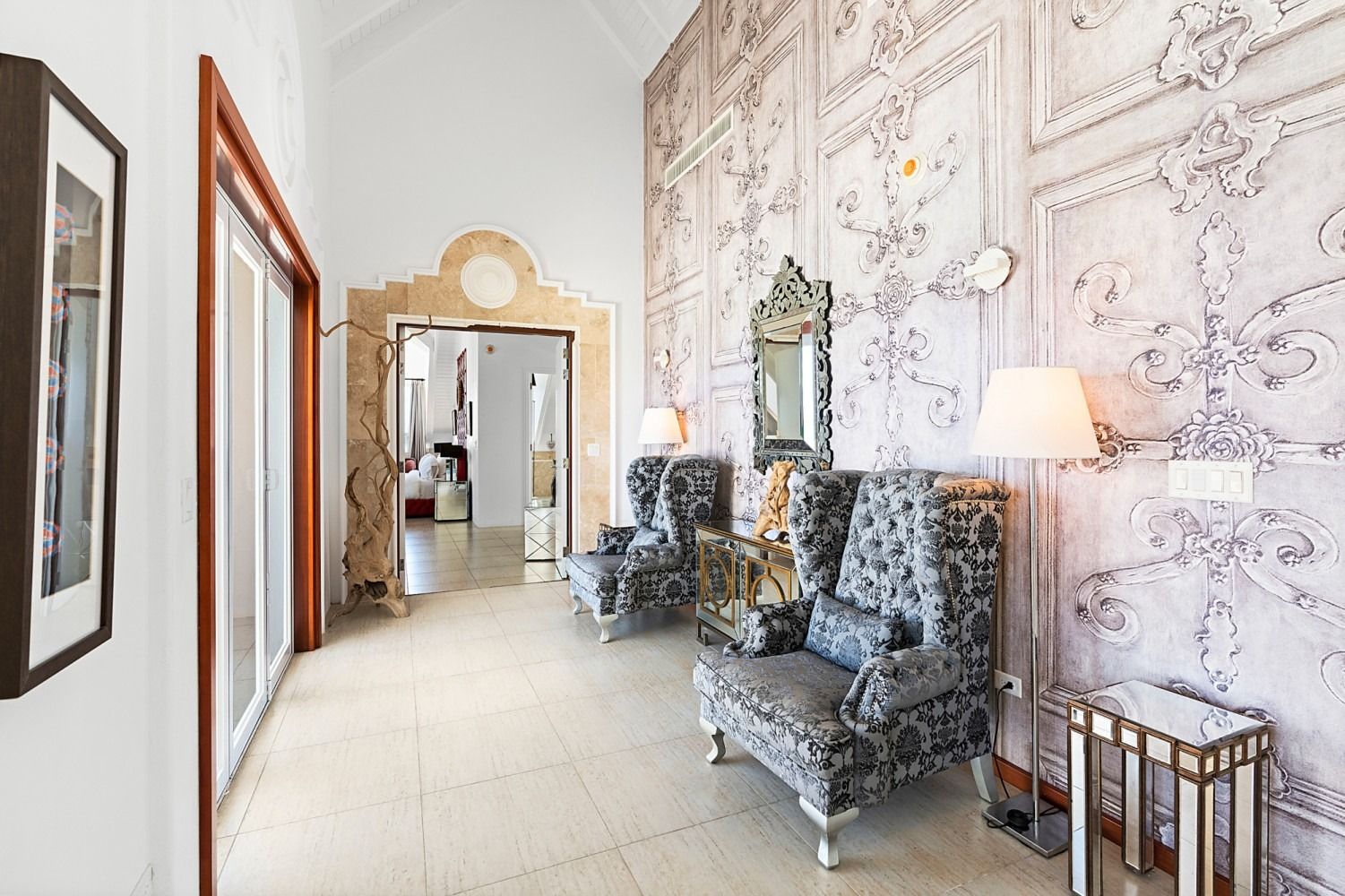 Elegant hallway with ornate wallpaper, two patterned armchairs, a mirror, and a lamp.