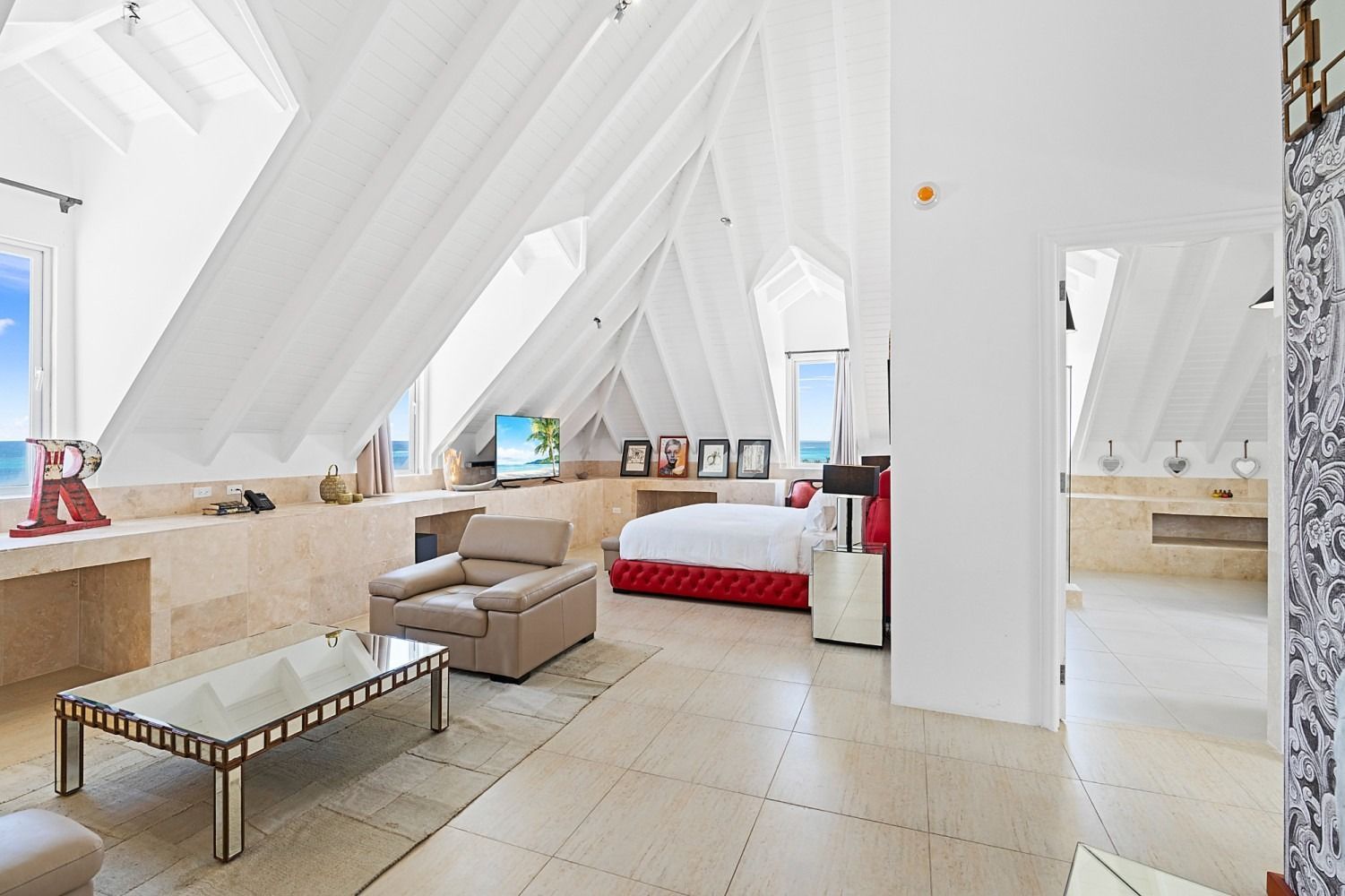 Bright, airy bedroom with high, draped ceiling, bed, seating area, and glimpses of the ocean.