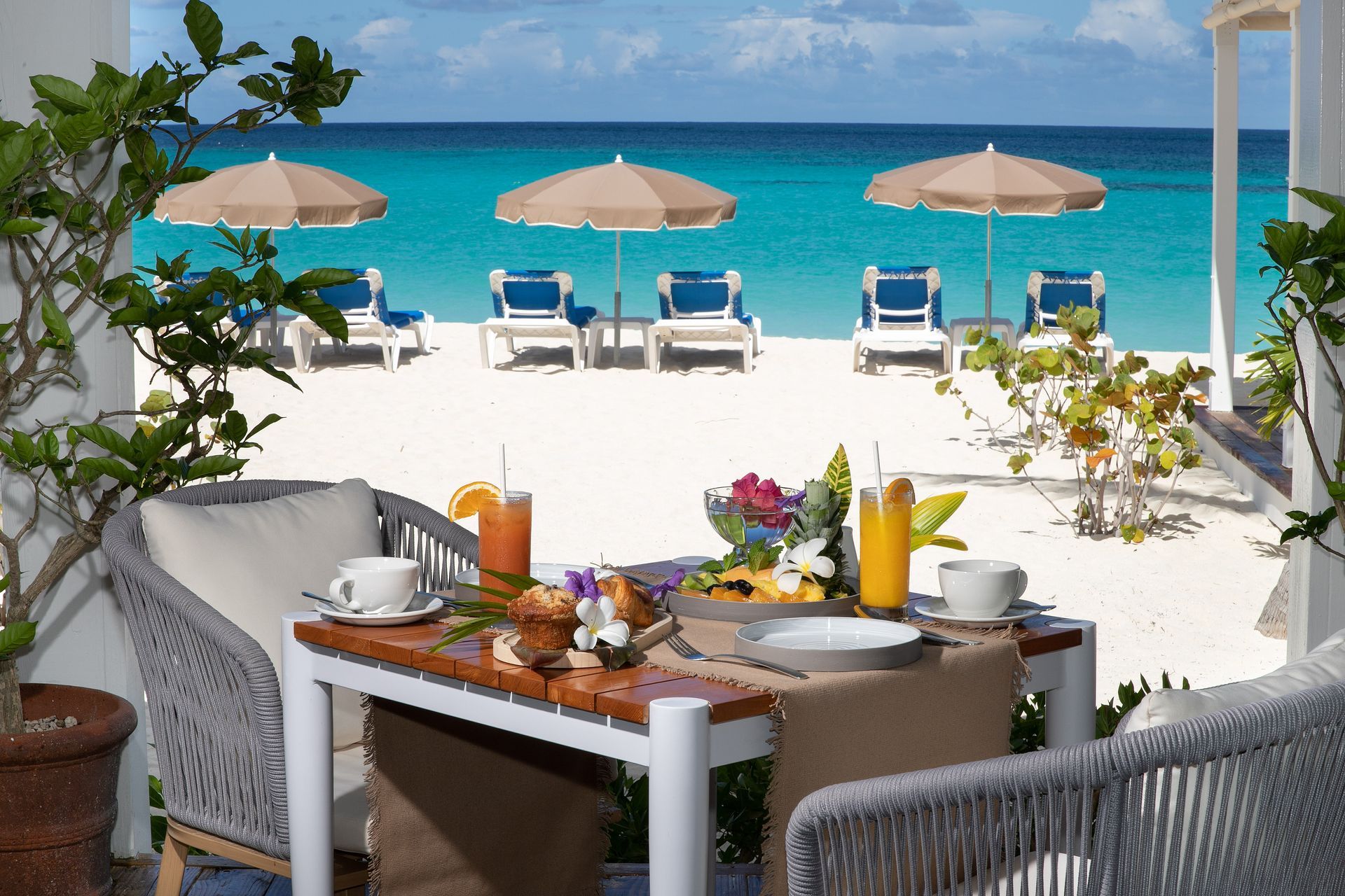 Beachfront table set for breakfast overlooking turquoise ocean, white sand, lounge chairs, and umbrellas