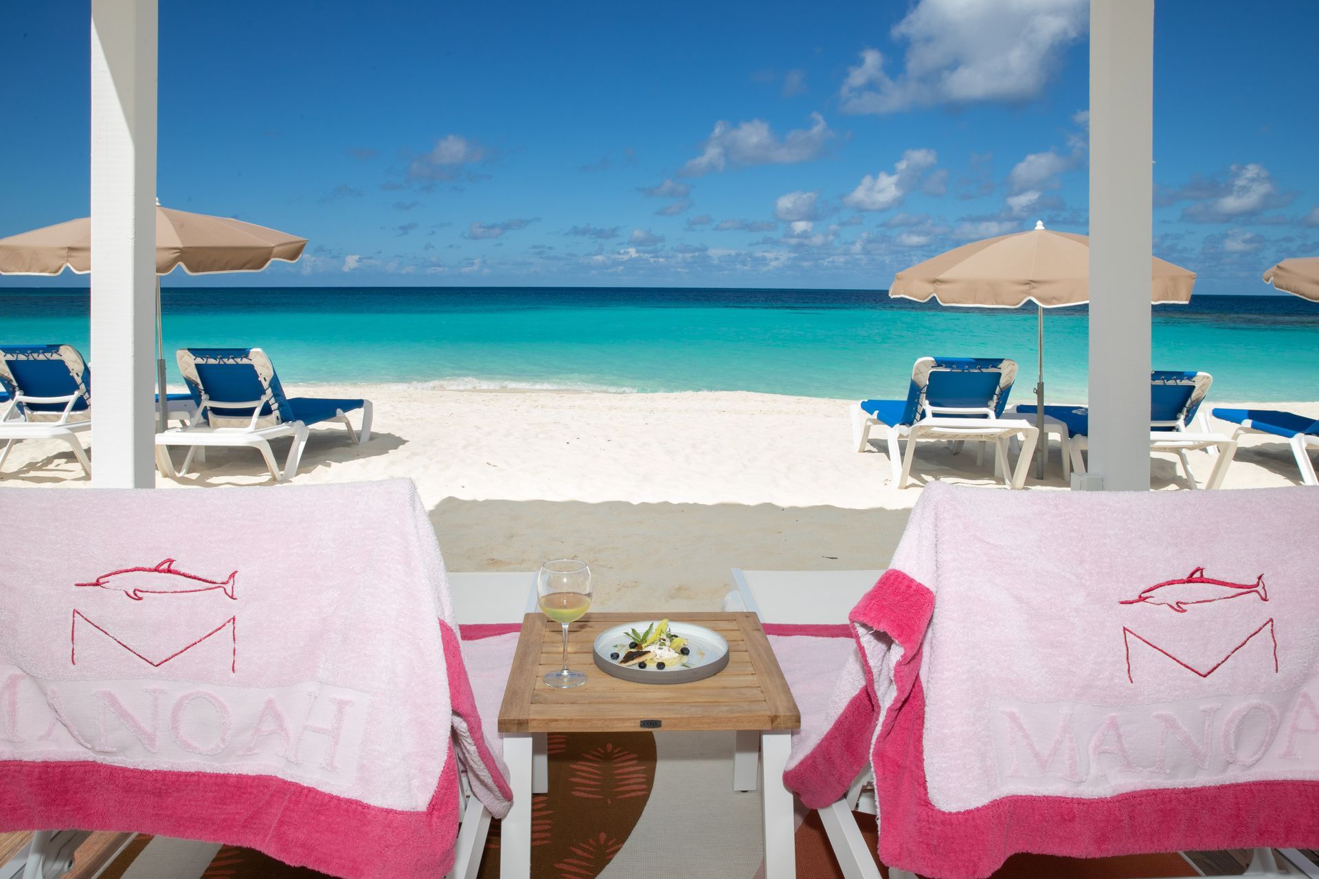 Beachfront cabana with lounge chairs, pink towels, and a turquoise ocean under a blue sky.