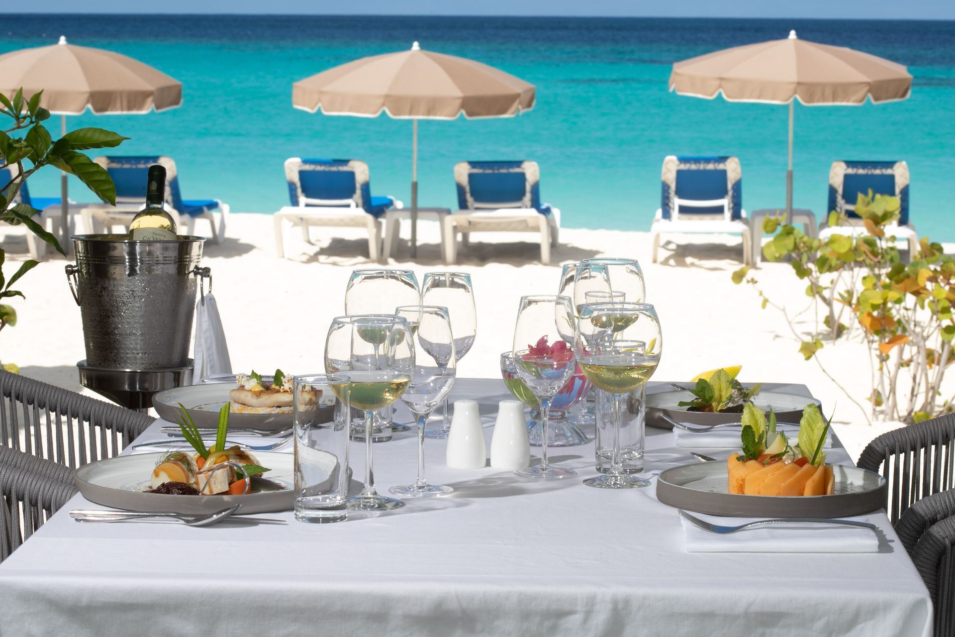 Beachfront dining table set with wine glasses, flowers, and white umbrellas overlooking blue water