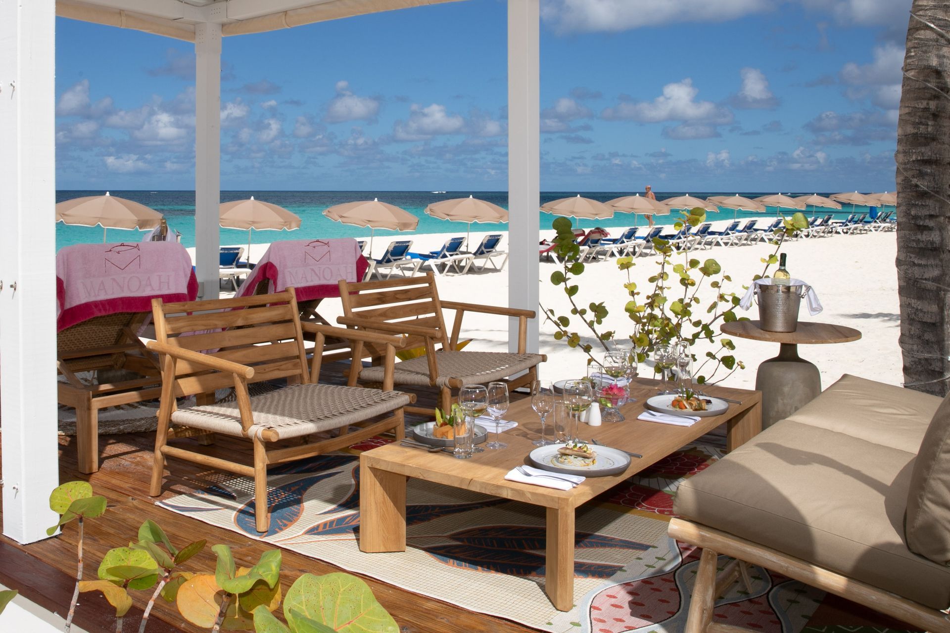 Beachside lounge with wooden seating, white sand, blue sea, and umbrellas under a sunny sky