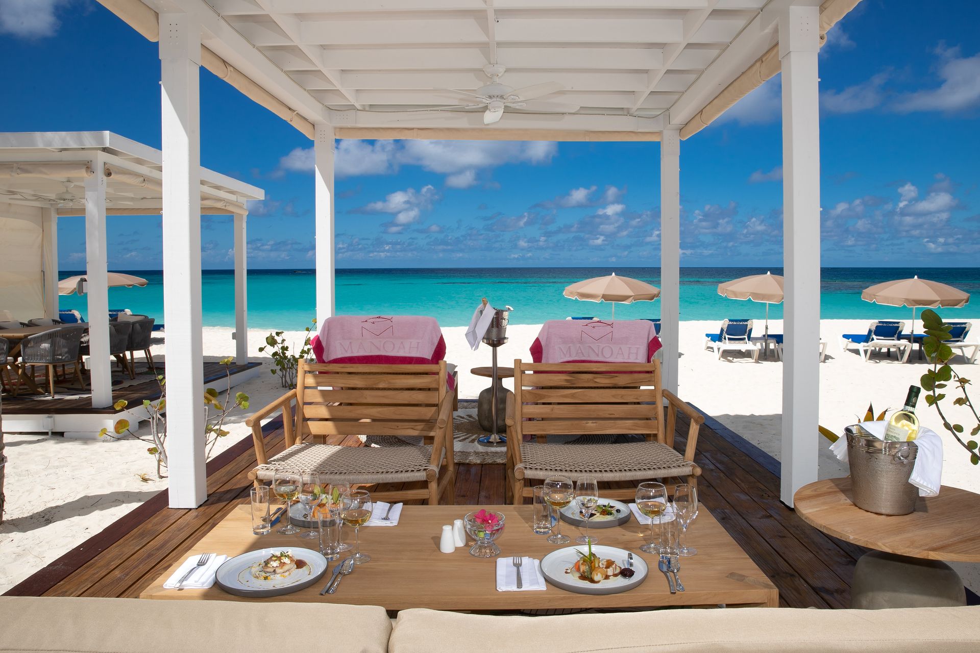 Beachside cabana with loungers, table setting, and turquoise ocean under a blue sky.