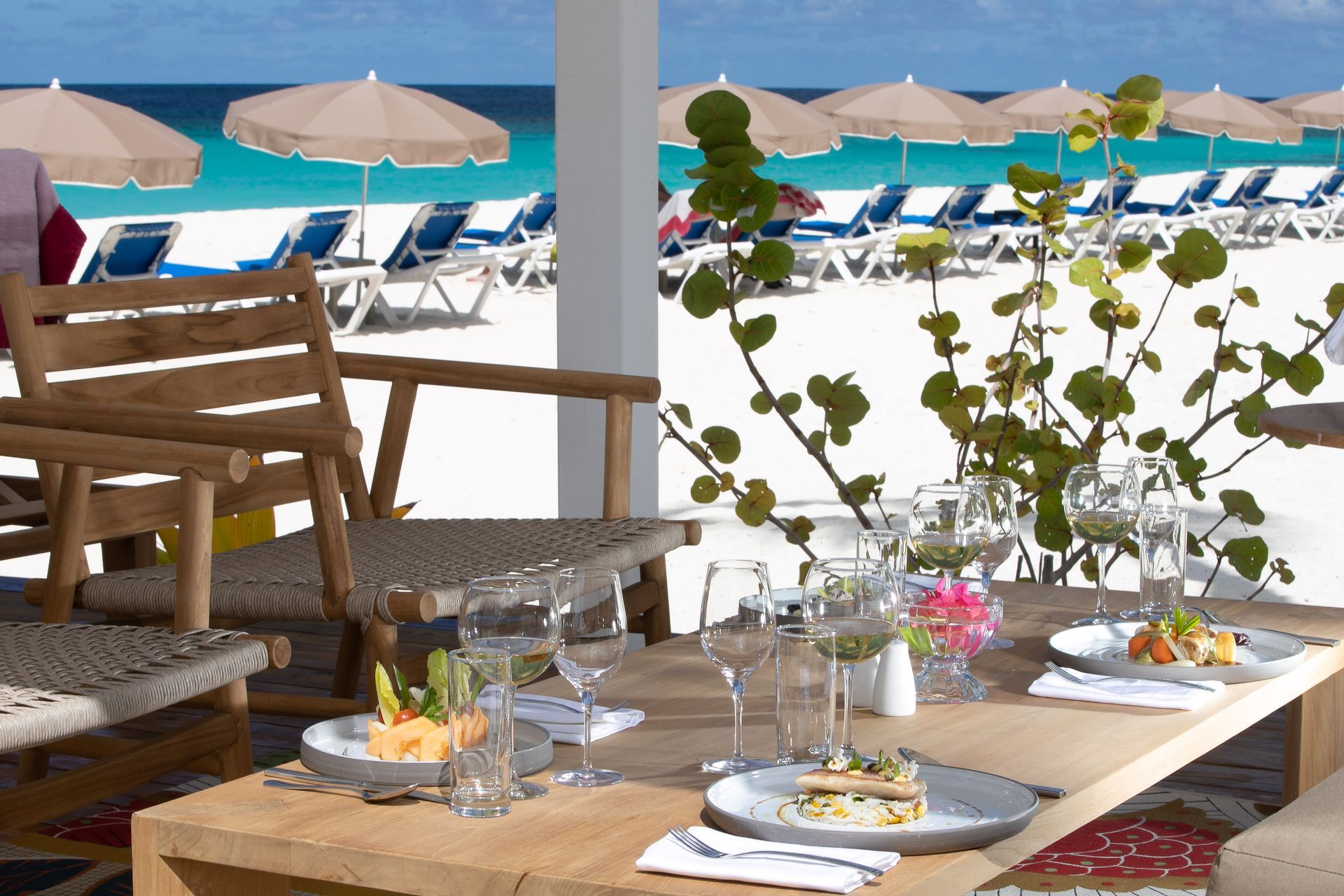 Beachside dining table with breakfast plates, wine glasses, and chairs overlooking lounge chairs and umbrellas by the sea