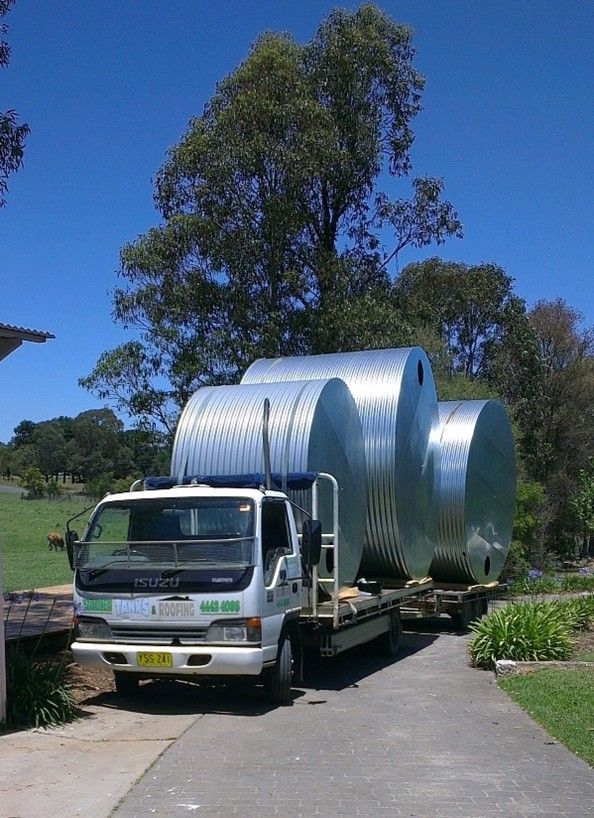 White flatbed truck carrying a large silver metal tank on a driveway with trees and blue sky behind White flatbed truck carrying a large silver metal tank on a driveway with trees and blue sky behind