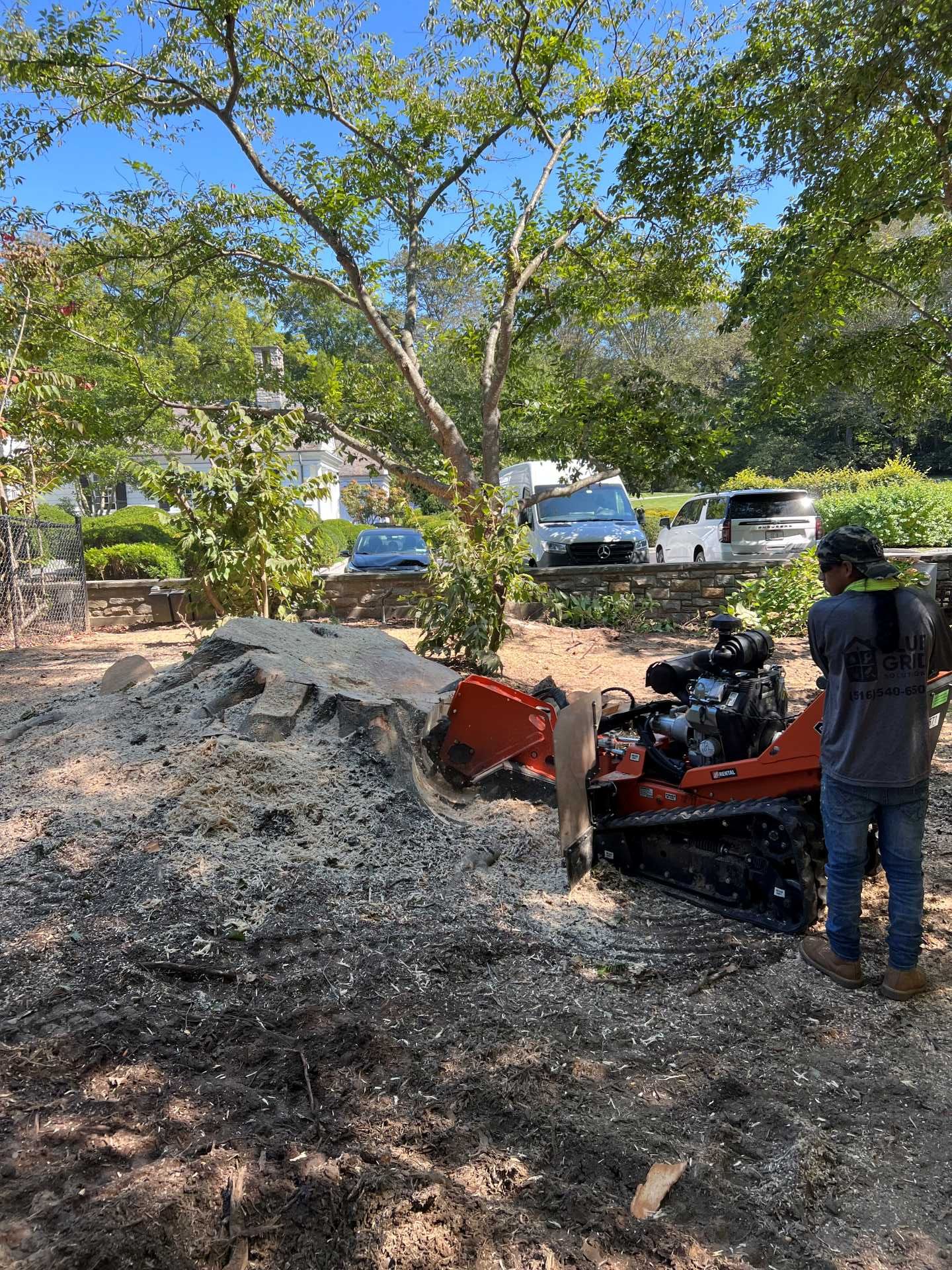 Man operating a stump grinder; dark debris pile, cars and trees in the background.