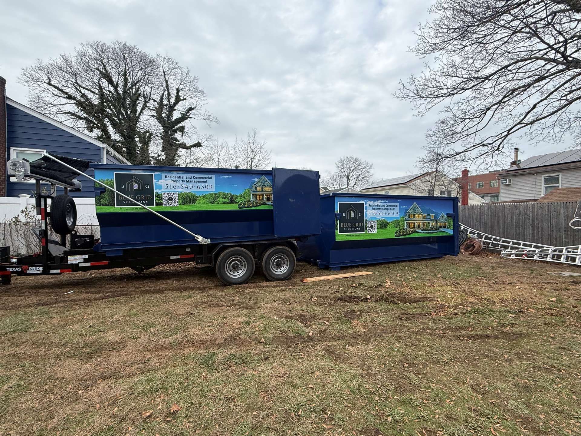 Blue dumpster trailer in a yard with house and fence in background.