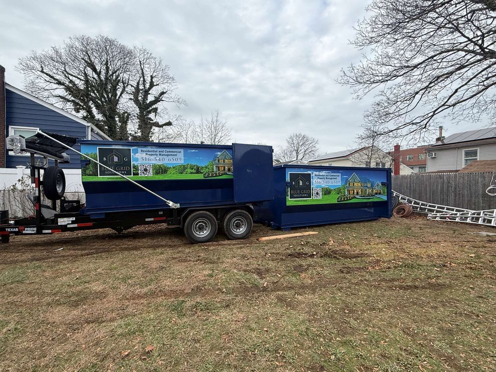 Blue dumpster trailer on a grassy lawn; houses and bare trees in the background.