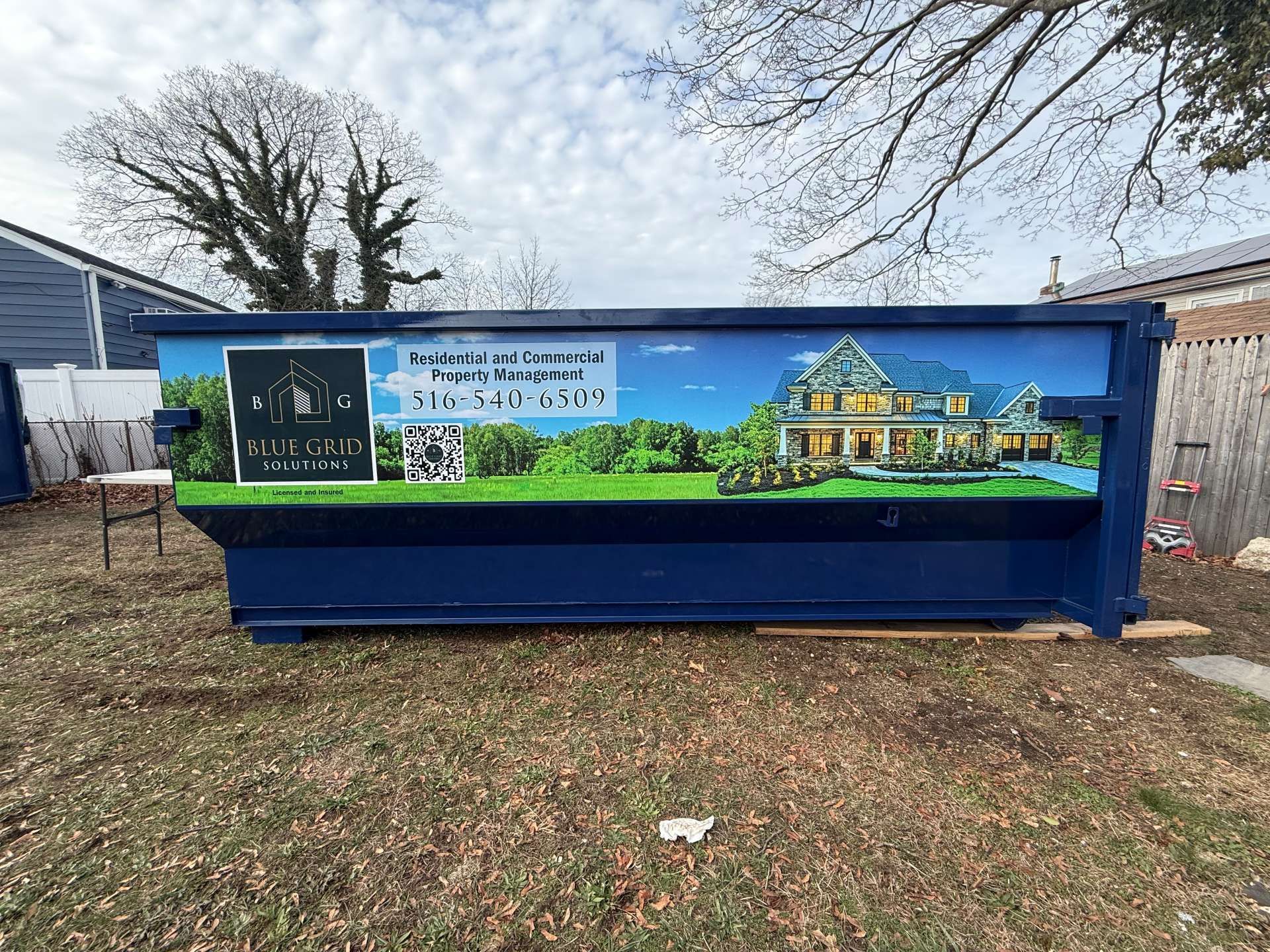 Blue dumpster with a mural of a house and trees in a backyard.