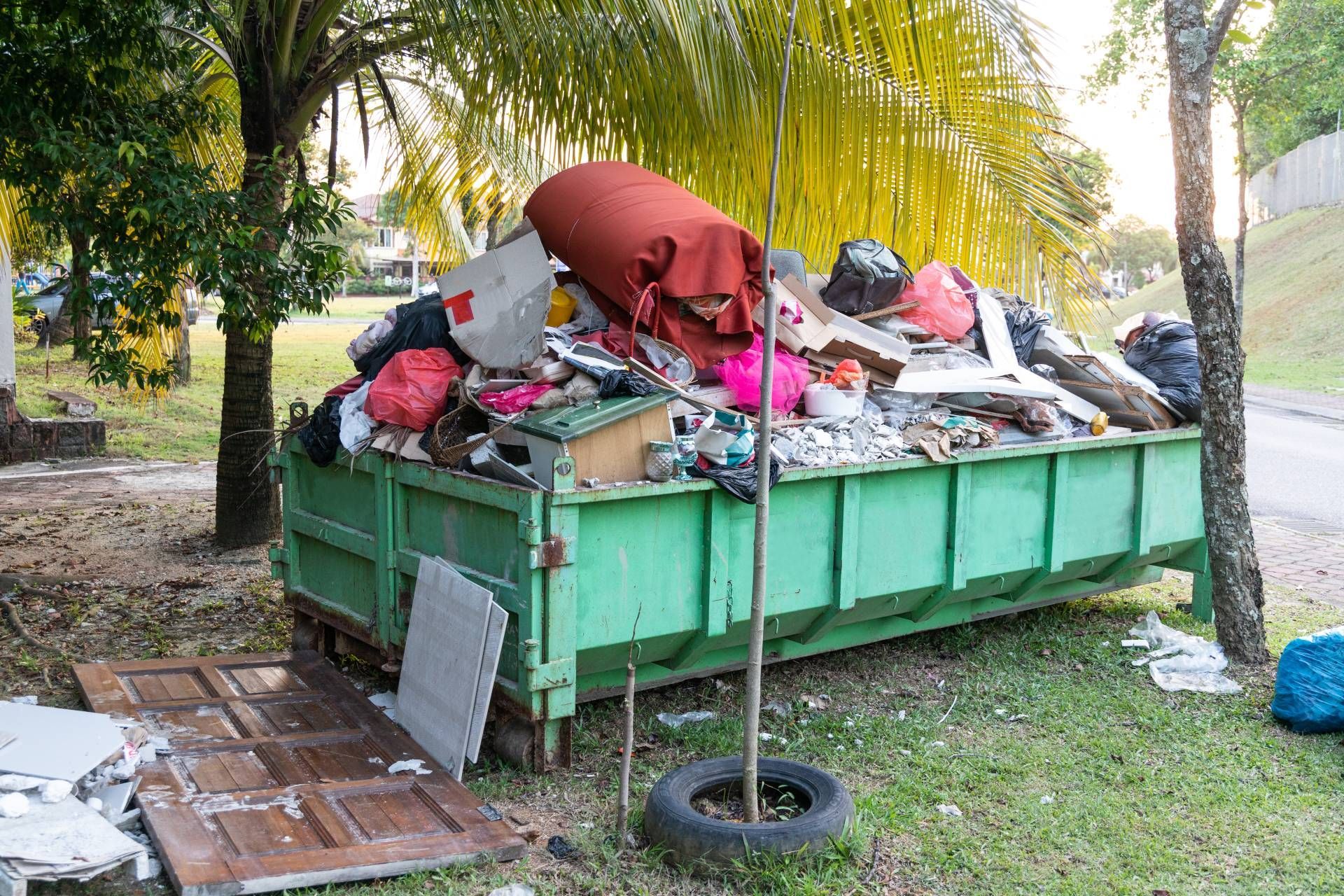 Green dumpster overflowing with trash near a road and trees. Old doors and debris are scattered nearby.
