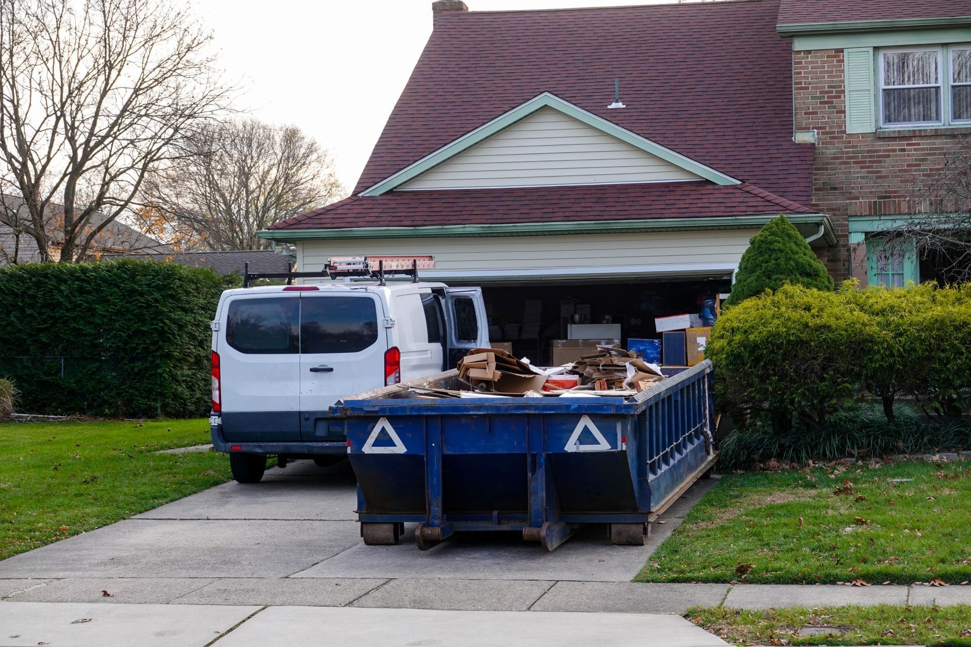 White van and blue dumpster in driveway of house with open garage.