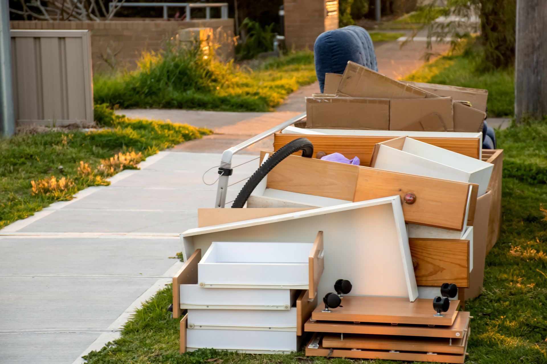 Heap of discarded furniture, including drawers and boxes, piled on a grassy verge next to a sidewalk.