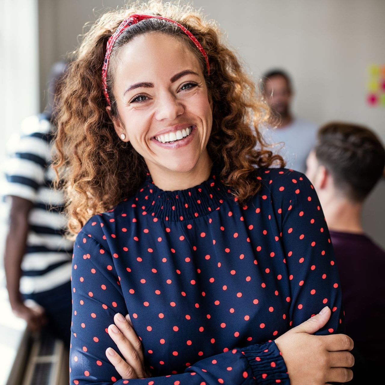 Woman with curly hair and a red headband smiles, arms crossed, in a casual office setting.