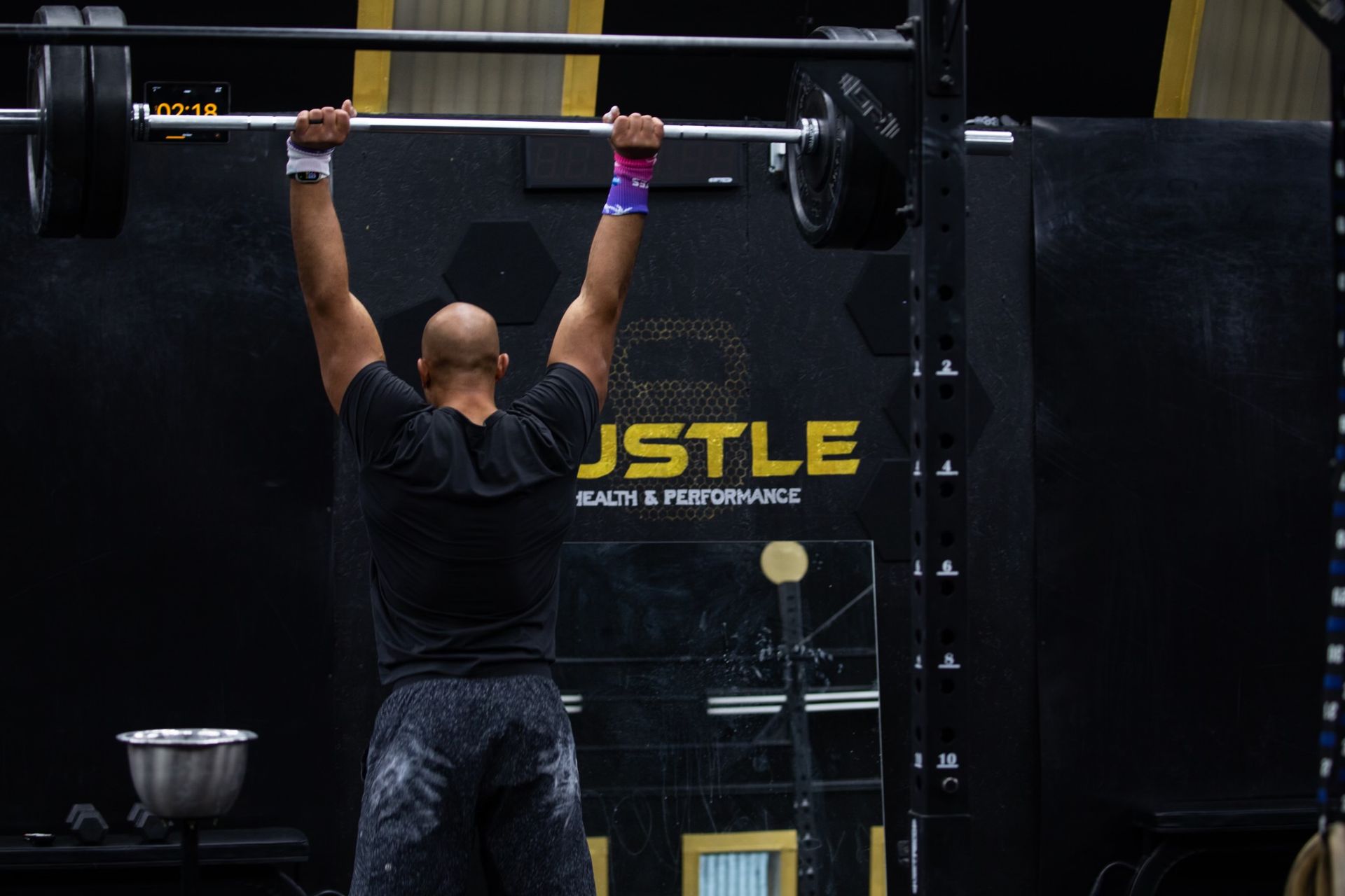 Person in black lifting a barbell overhead in a gym with JSTLE signage behind them