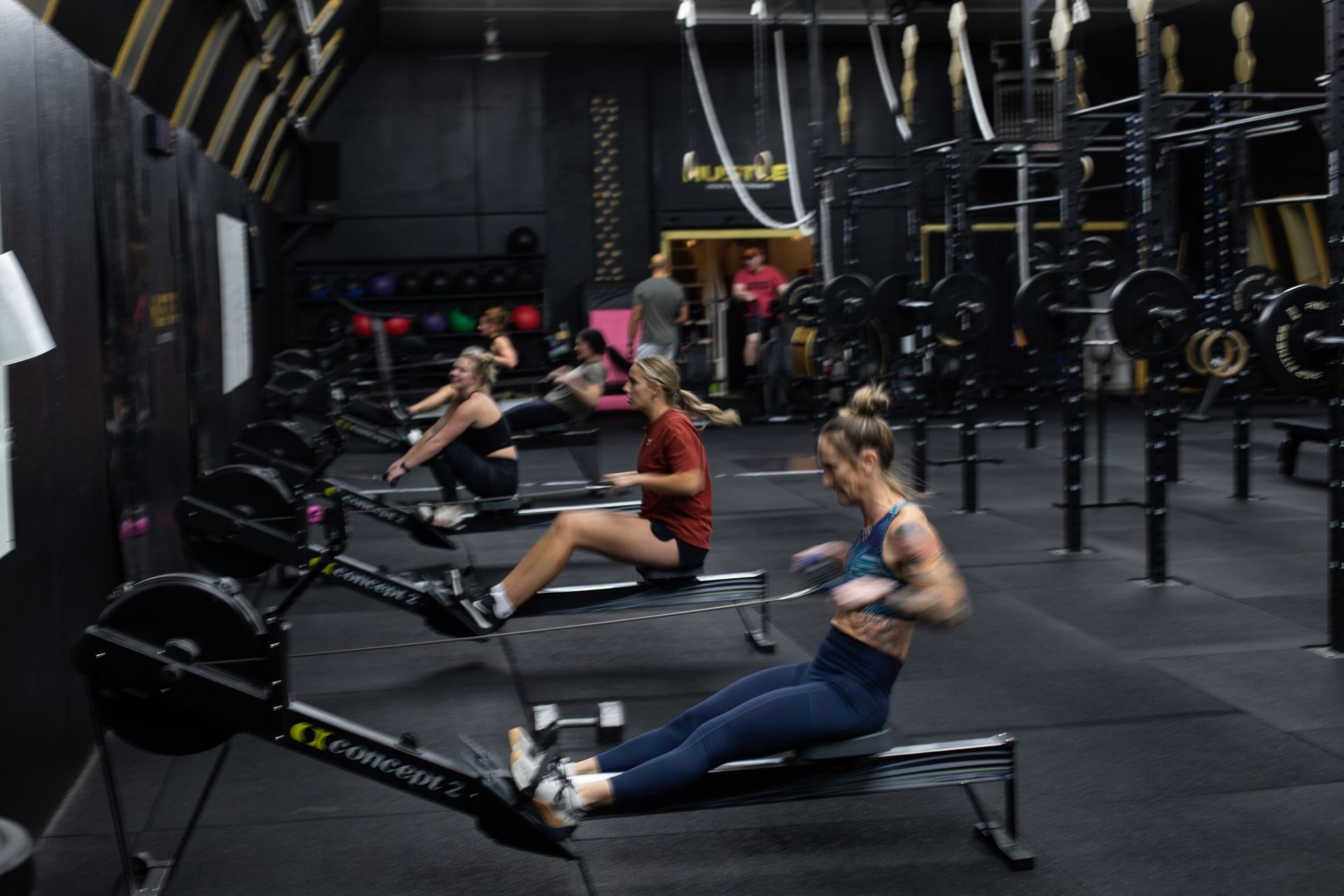 People rowing on erg machines in a dark indoor gym with suspension straps and weights