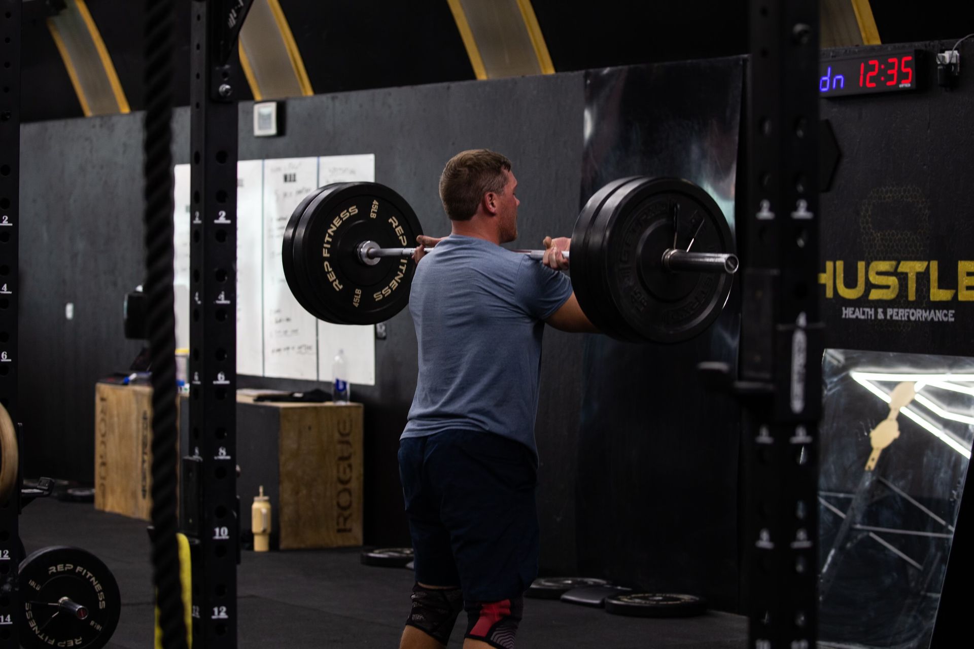 Person lifting a barbell in a gym with black equipment and a digital timer in the background