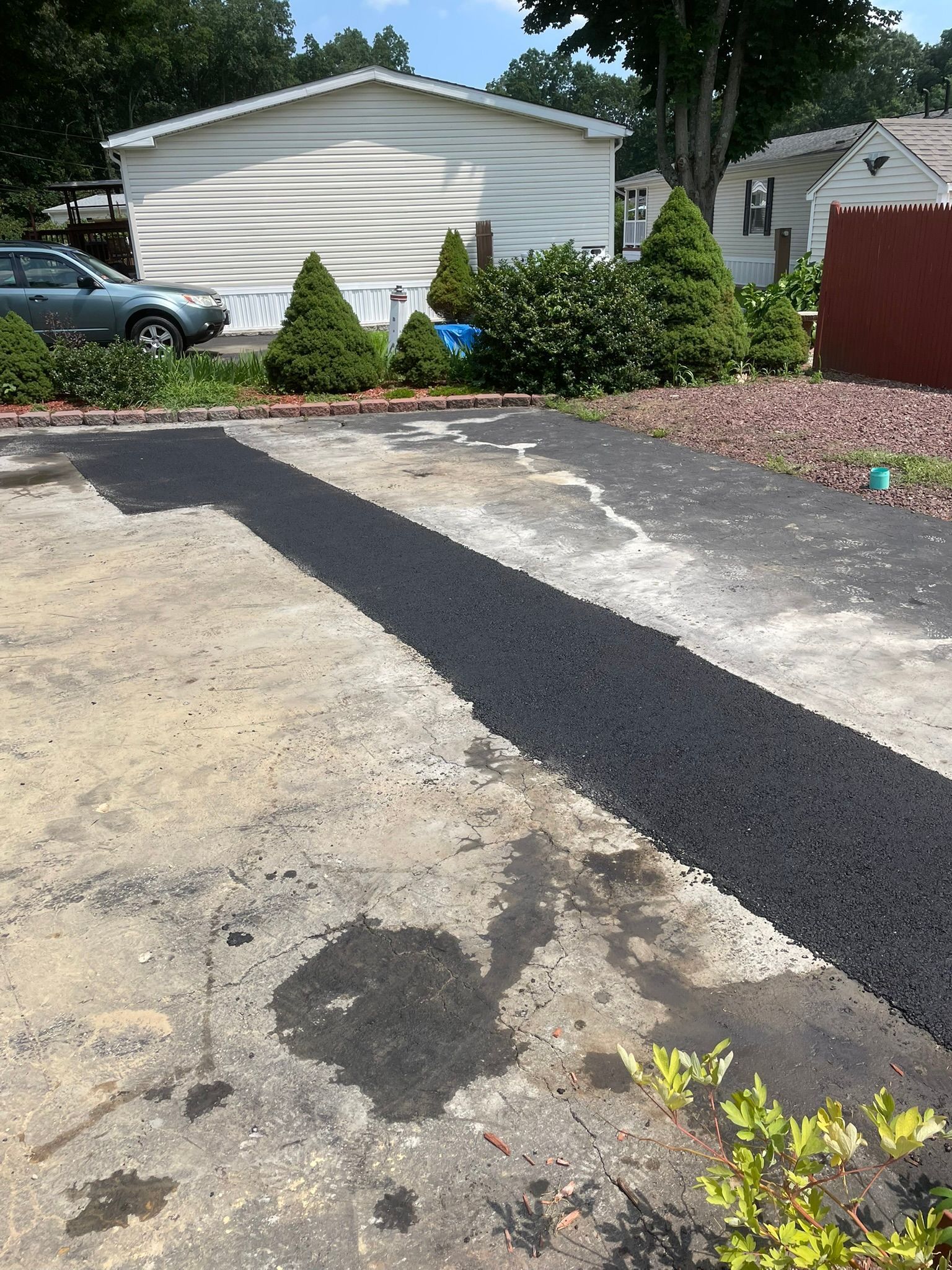 Driveway with a fresh black asphalt repair patch beside a white garage and parked cars.