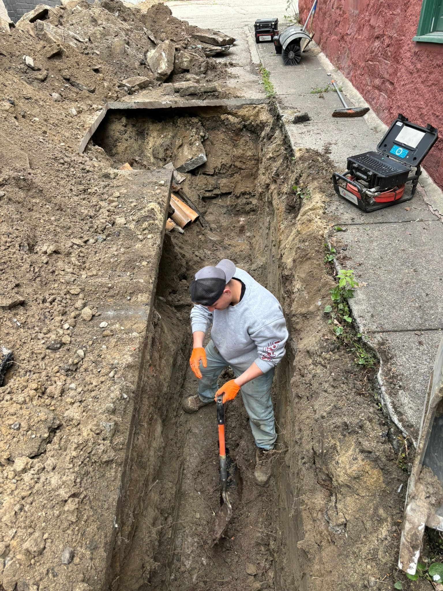 Worker digging a narrow trench beside a building, with excavator tracks and construction debris nearby