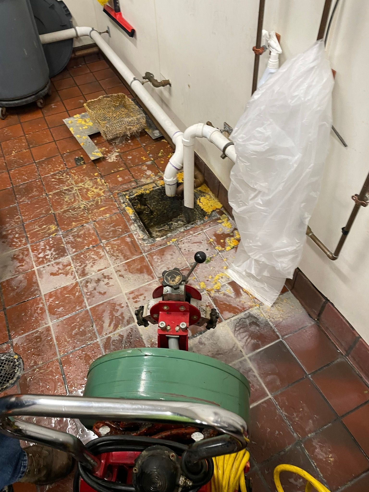 Red drain-cleaning machine in a tiled utility room beside exposed pipes and a plastic-covered wall.
