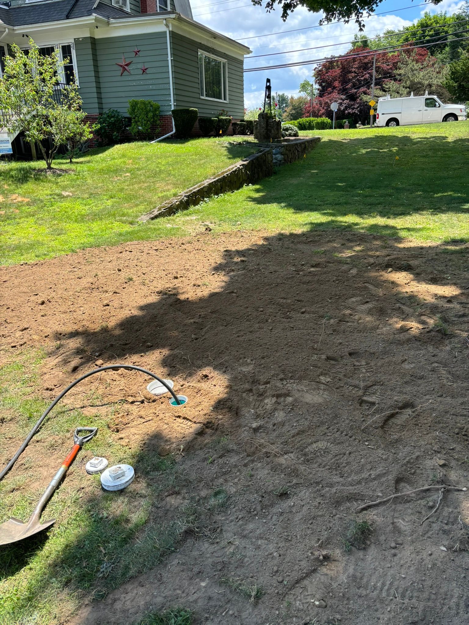 Residential yard with bare dirt patch, garden tools, and a house in the background on a sunny day