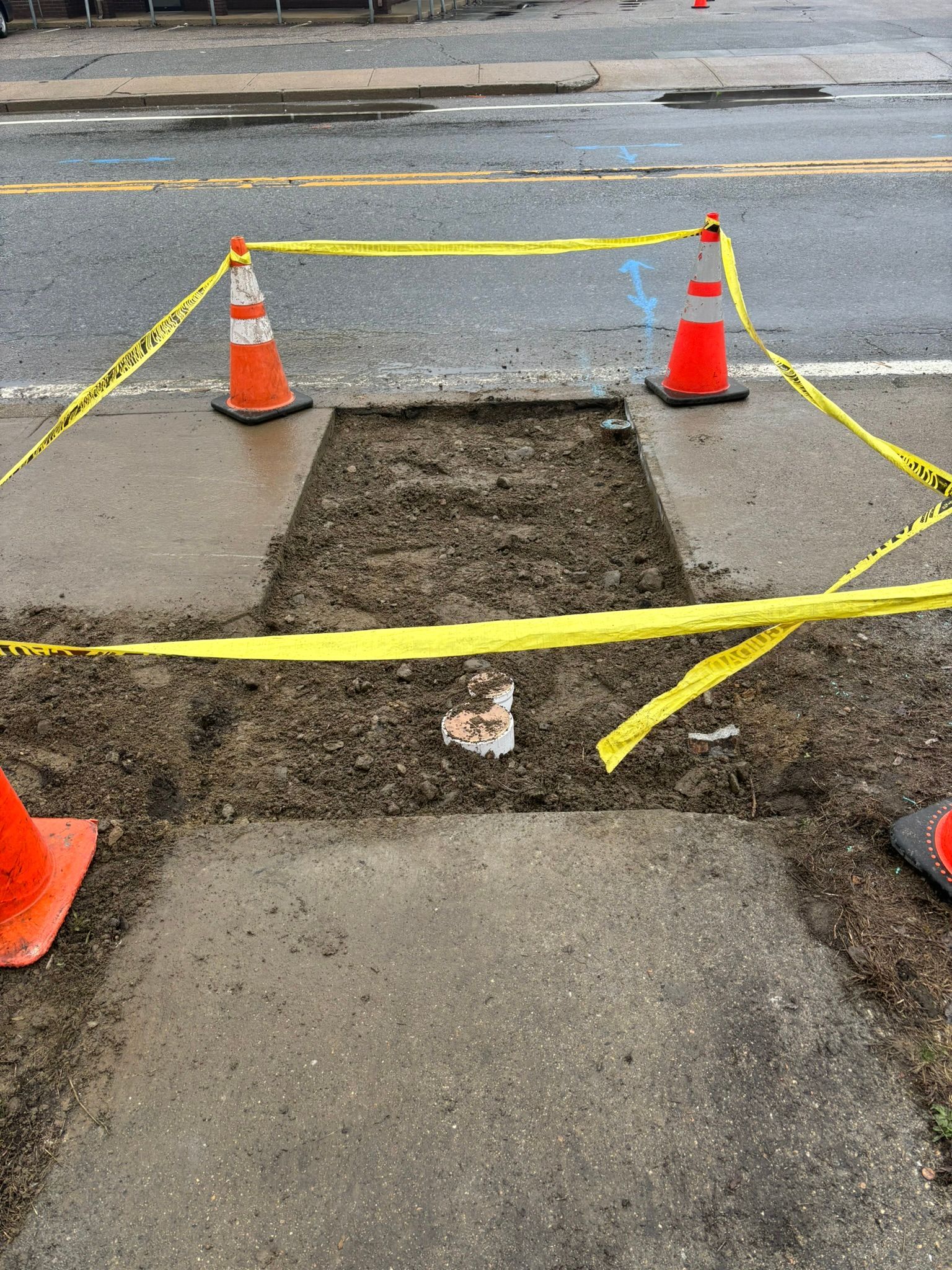 Blocked sidewalk repair area marked by orange cones and yellow caution tape, with a rough excavation in the pavement