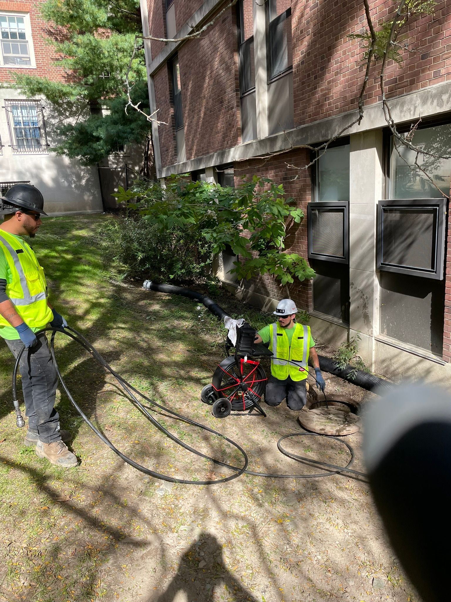Two utility workers in safety vests inspect underground cables beside a brick building.