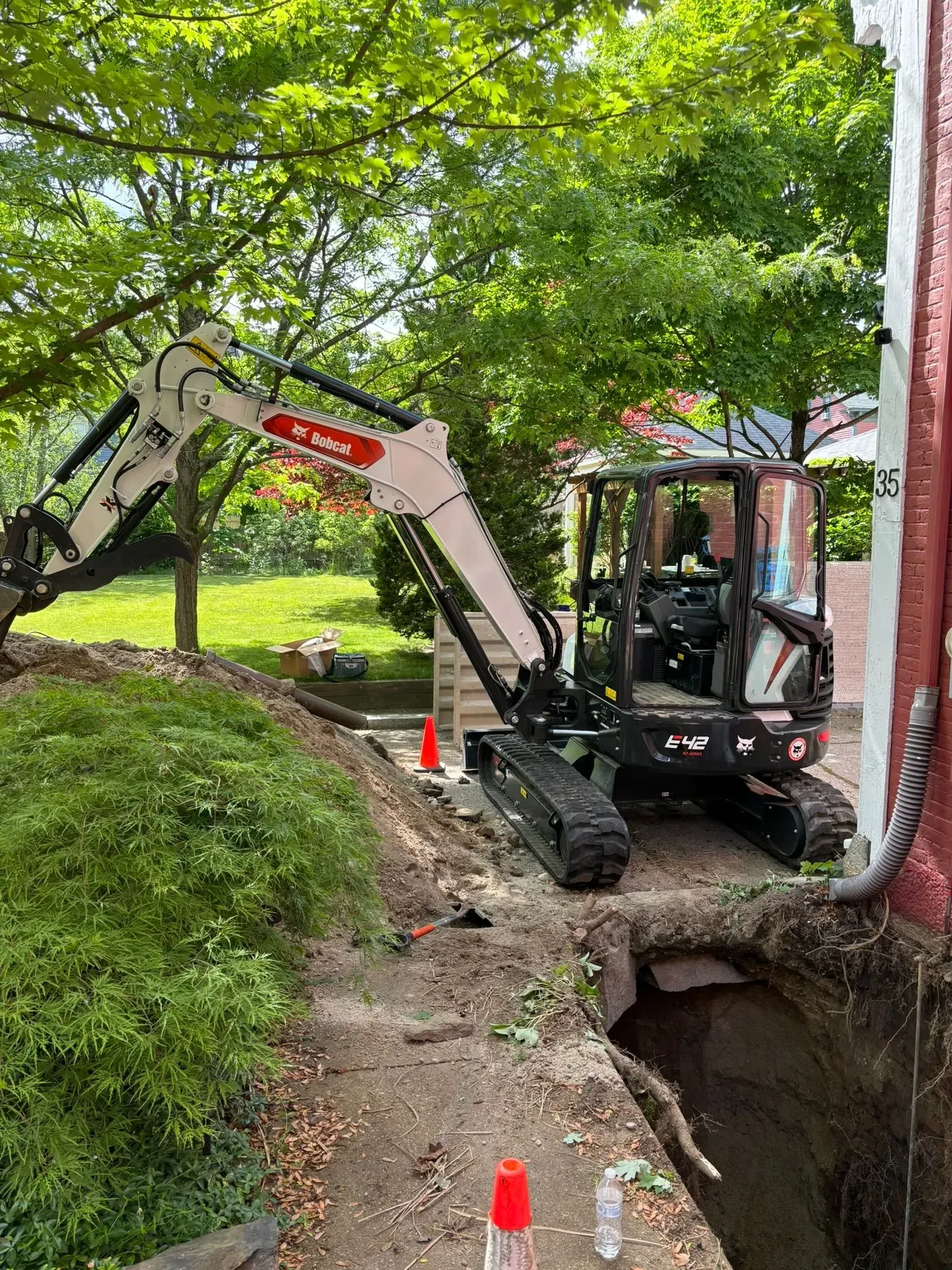 Excavator working beside a trench on a wooded path, with traffic cones marking the excavation area.