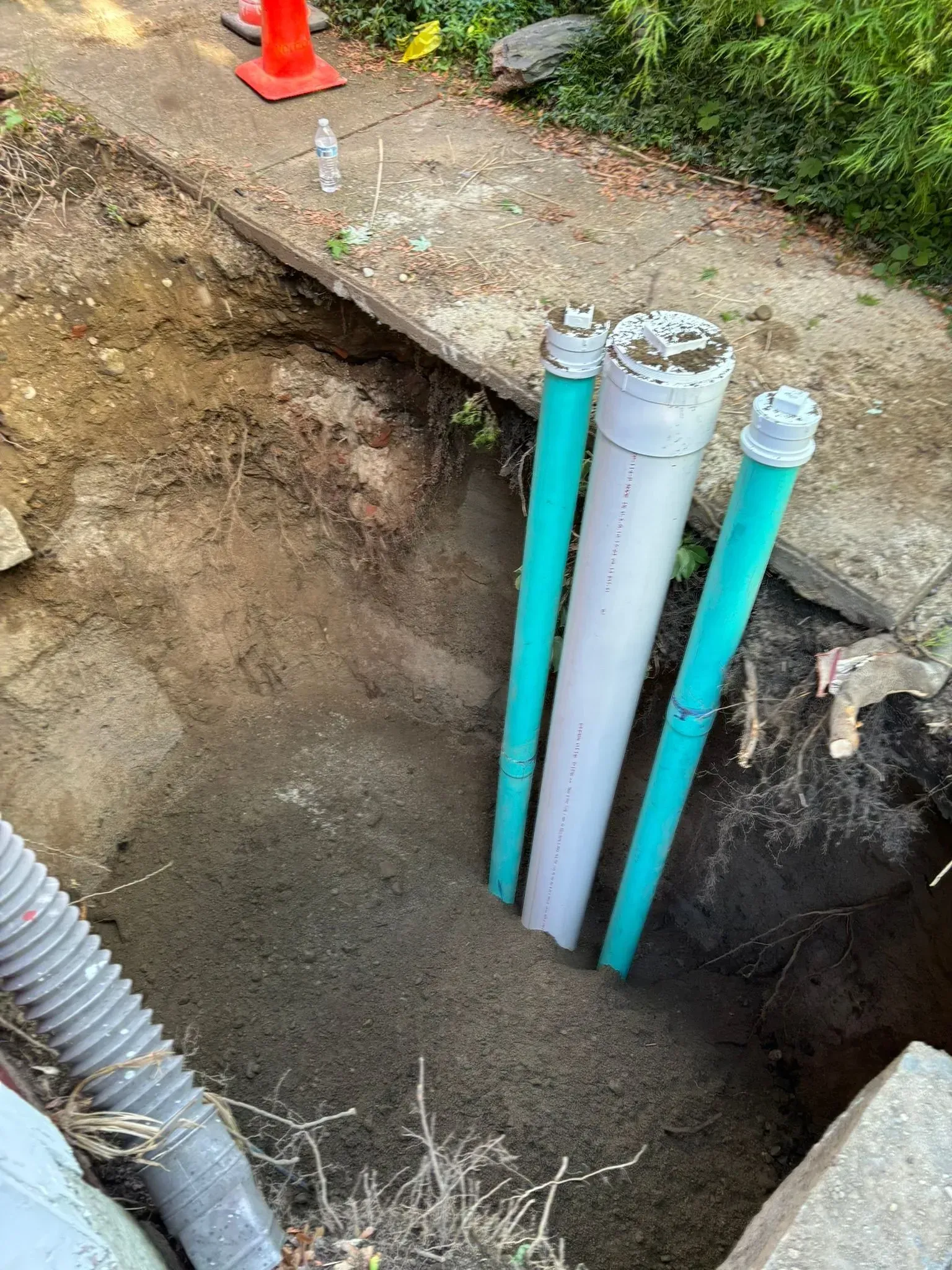Open utility trench with three vertical PVC pipes beside a roadside curb and orange cone.