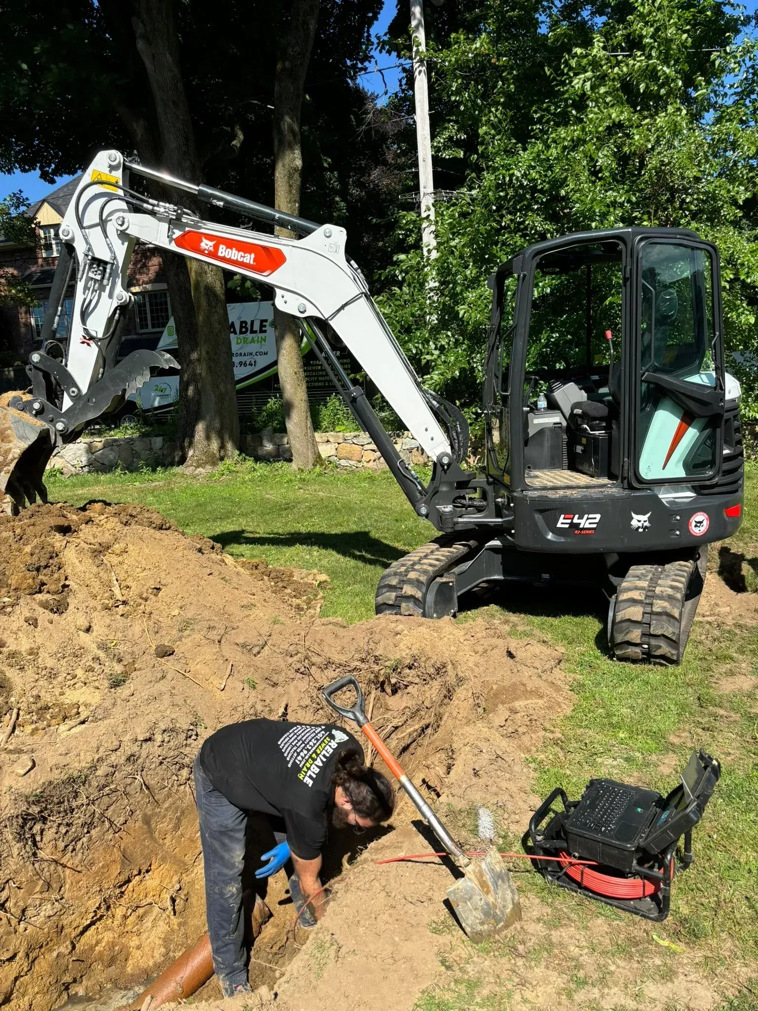 Worker digging near a small excavator at a wooded construction site, with tools and dirt pile nearby
