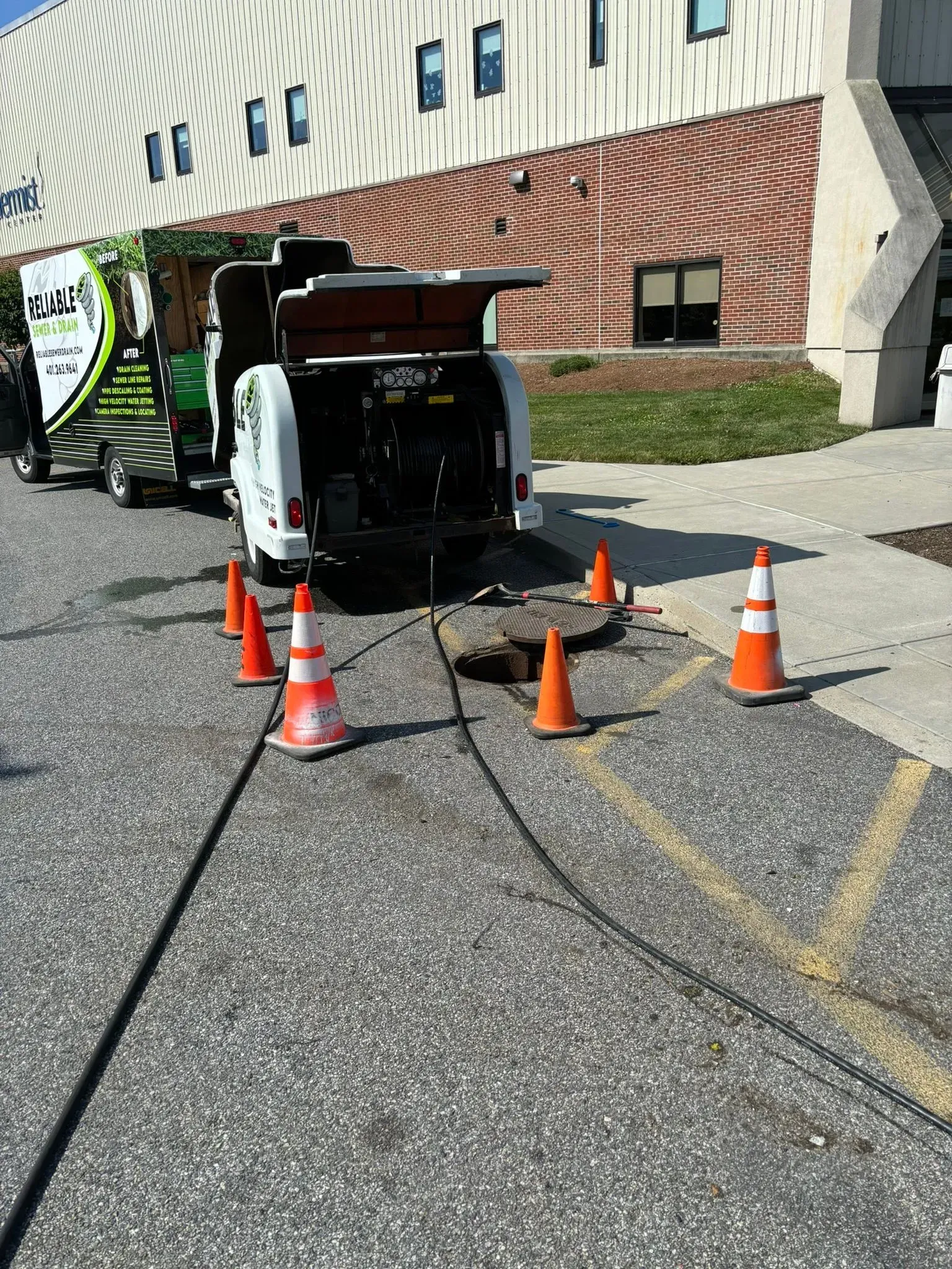 White utility truck with open rear door, orange cones, and cables in a parking lot beside a building.
