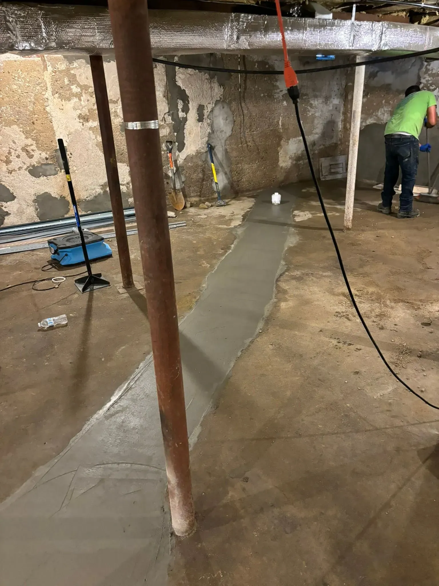 Basement construction site with a wet concrete channel, cables, and a worker in a green shirt near the wall