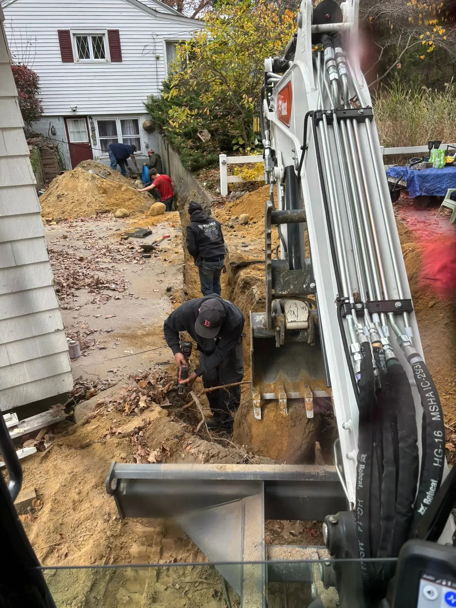 Workers dig a trench beside a house while an excavator arm lowers a metal beam on a muddy site.
