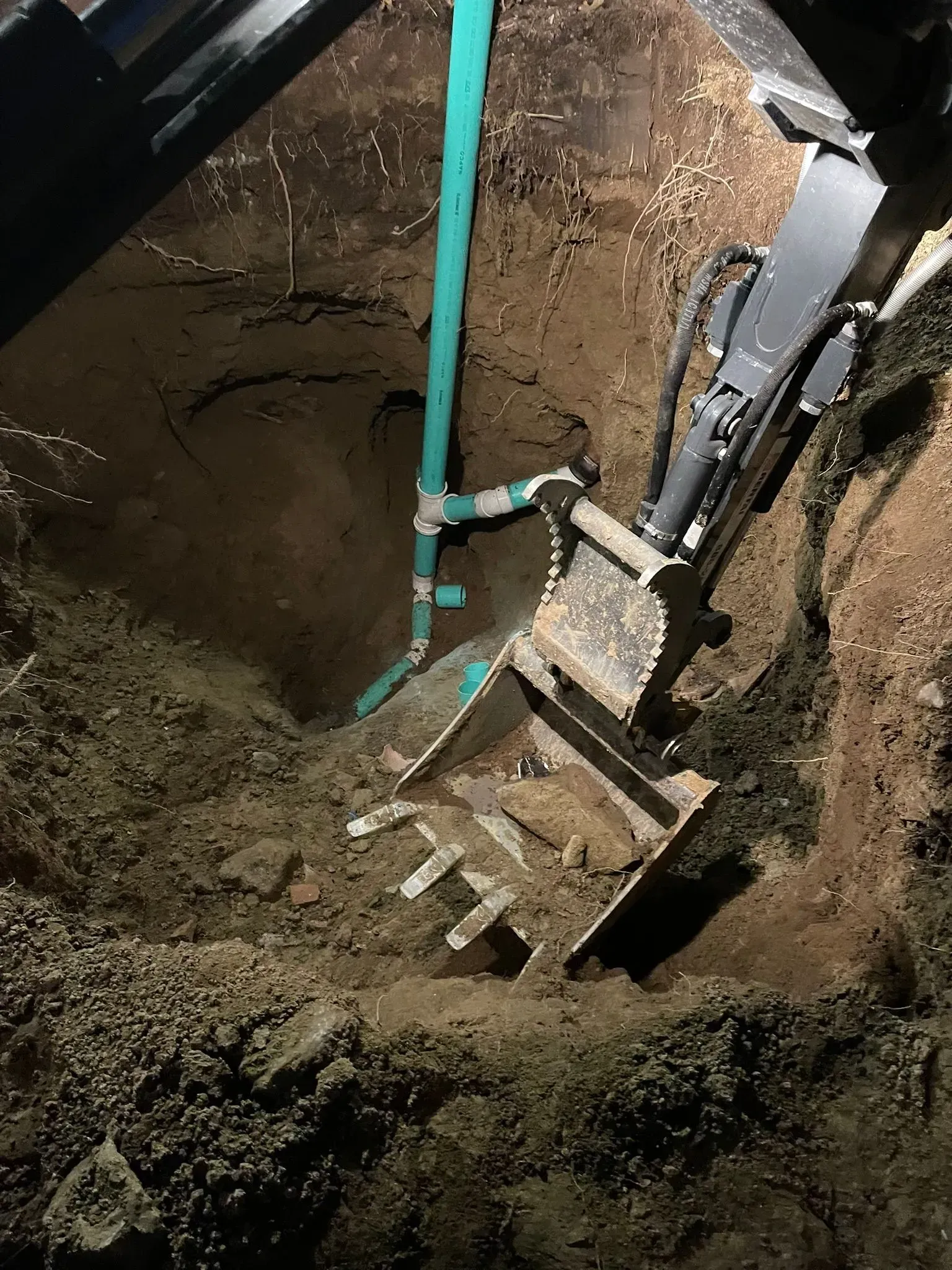 Excavator bucket digging a muddy trench beside a green pipe and soil wall