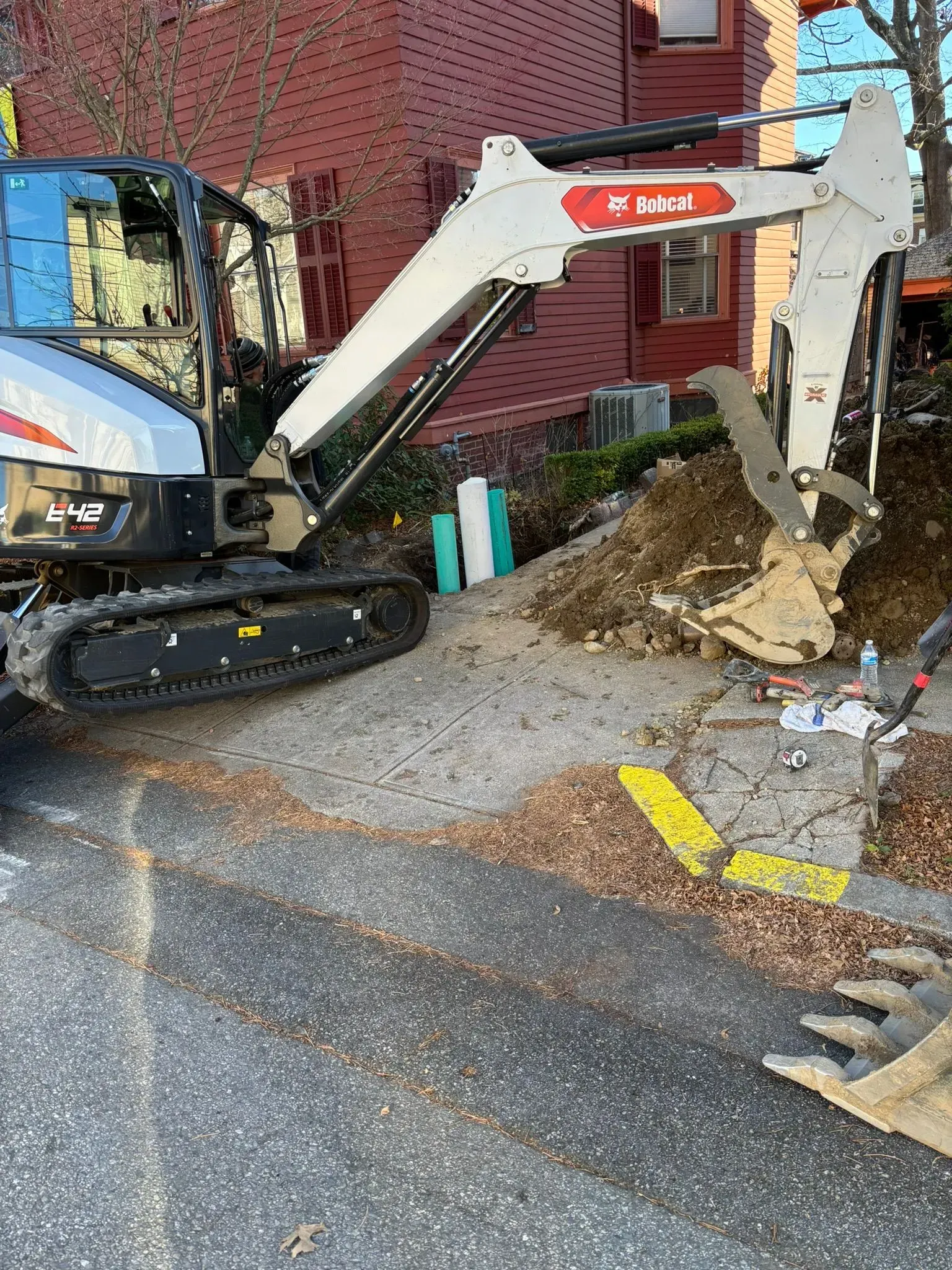 Mini excavator digging dirt beside a brick building on a street curb