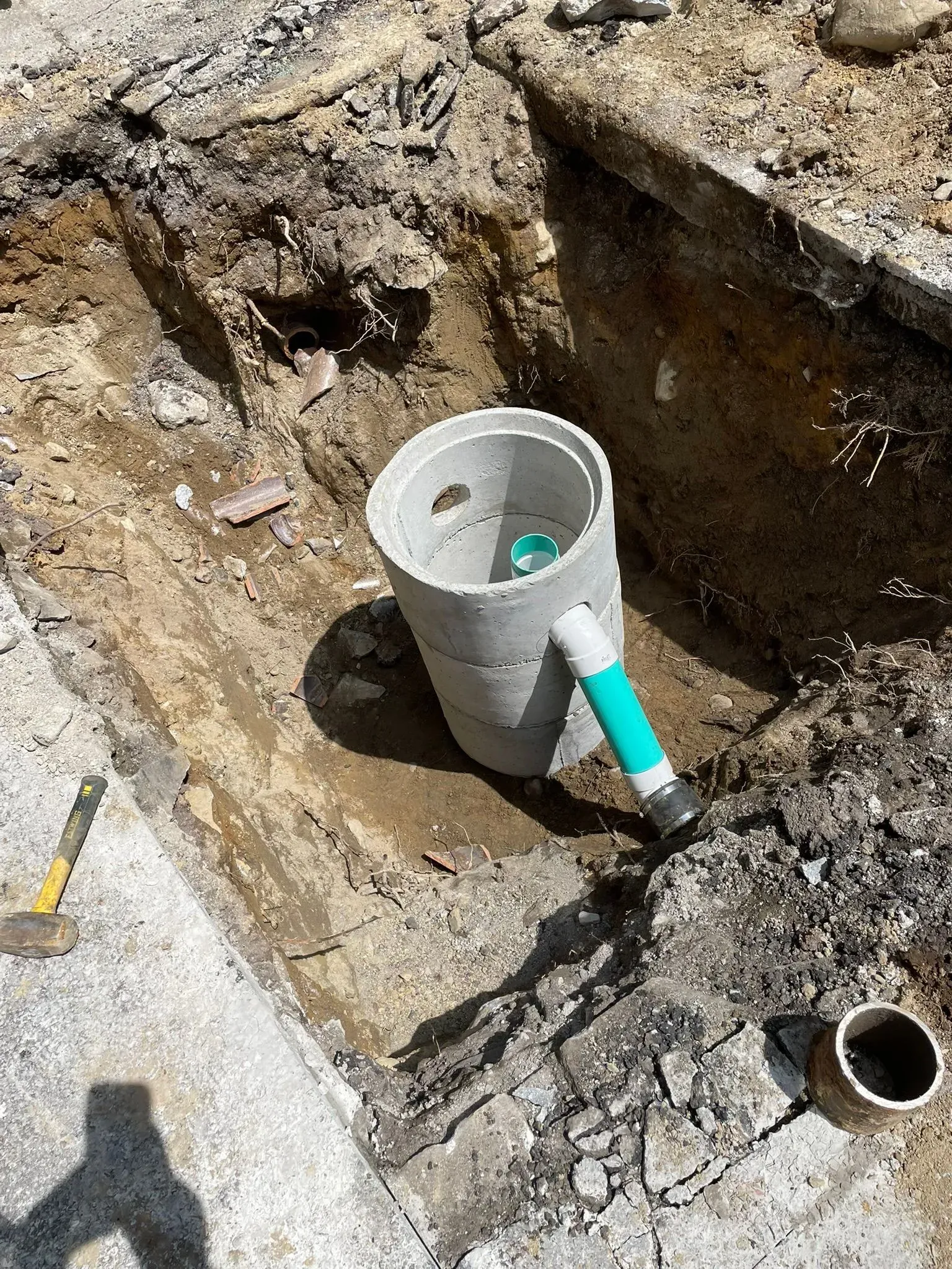 Excavation pit with a white PVC pipe and green fitting in dirt near a concrete edge